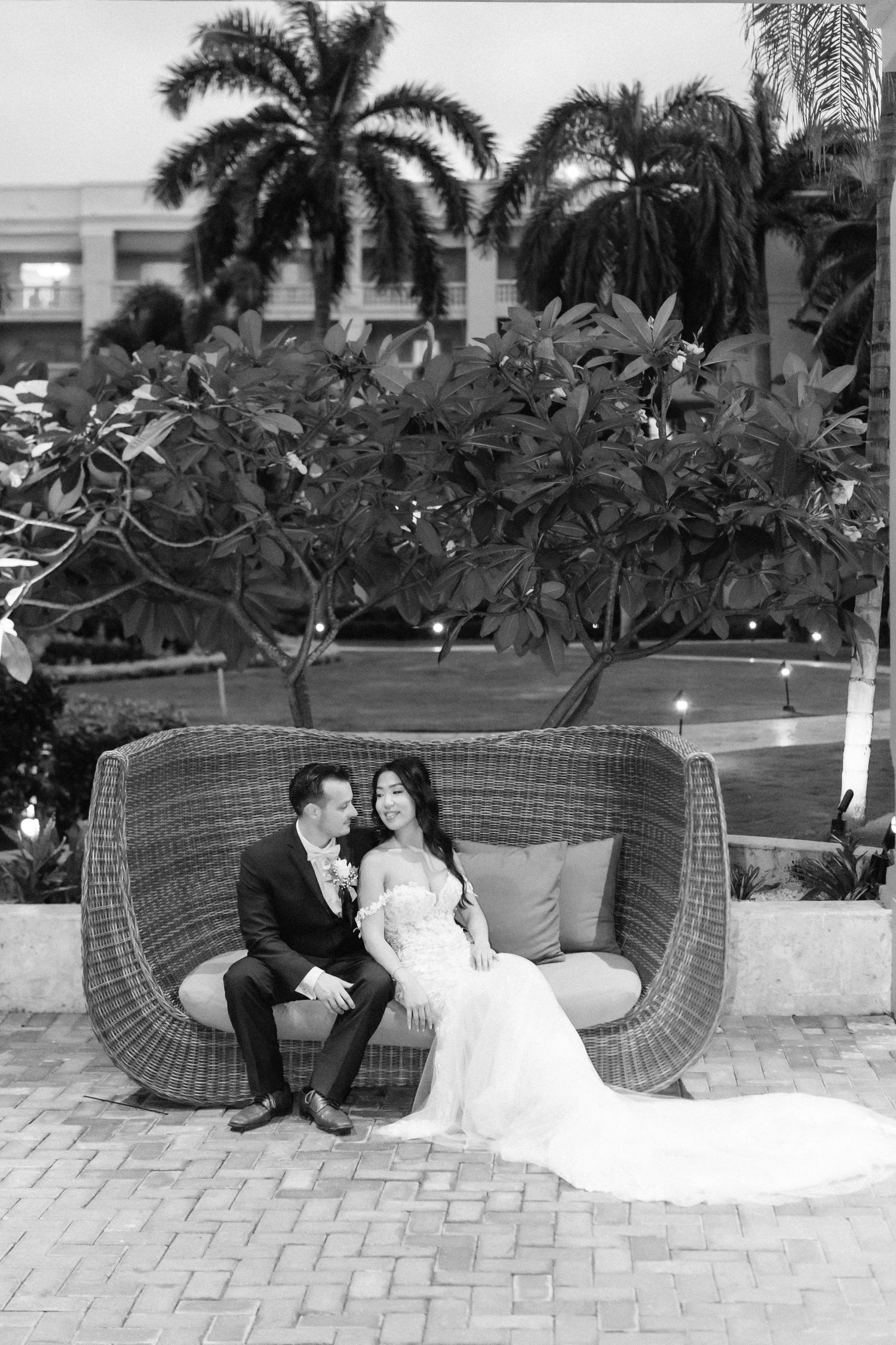 A romantic wedding couple sits in a woven outdoor lounge chair surrounded by tropical palm trees.