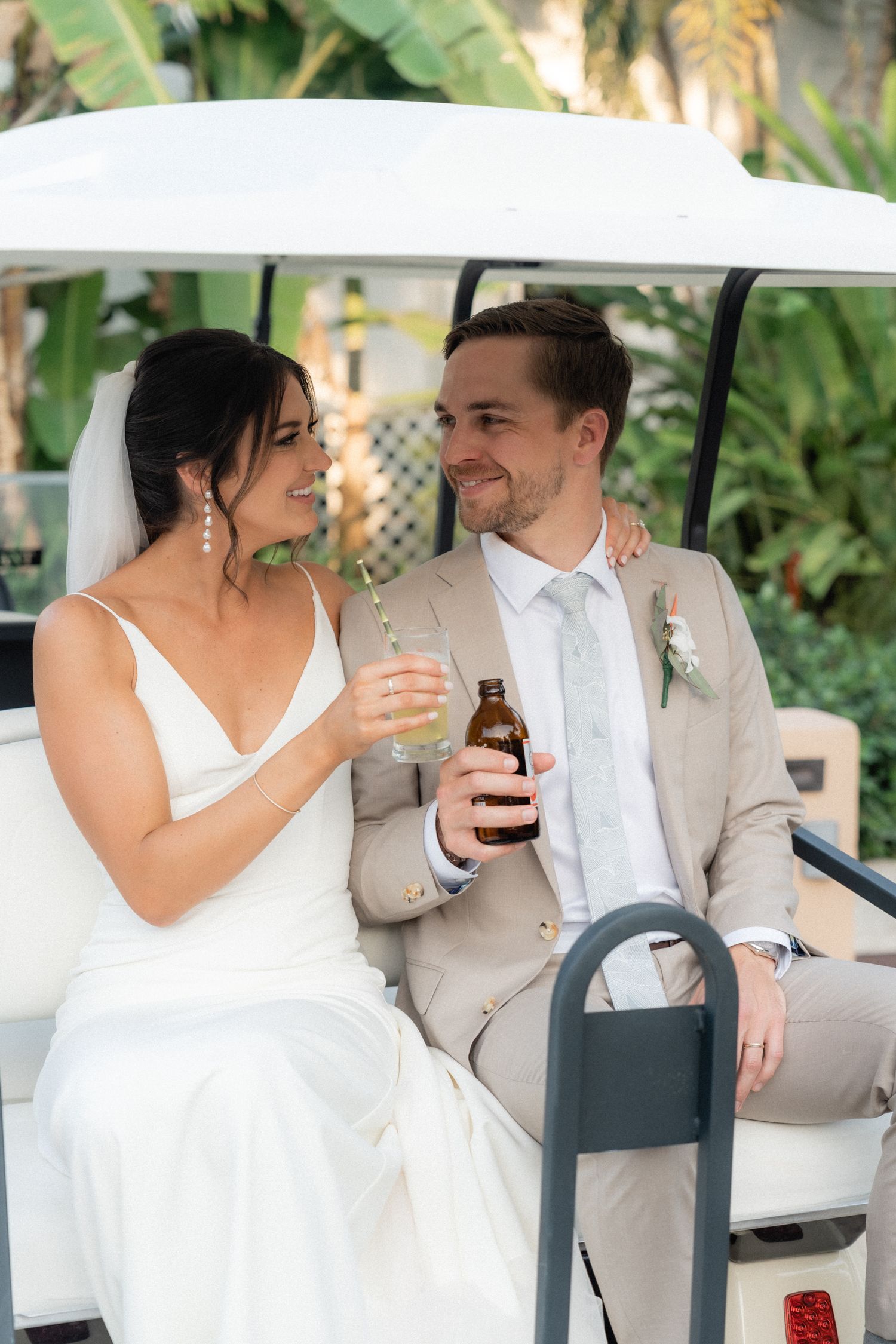 Newlyweds sharing drinks while riding in a golf cart through a tropical garden setting.