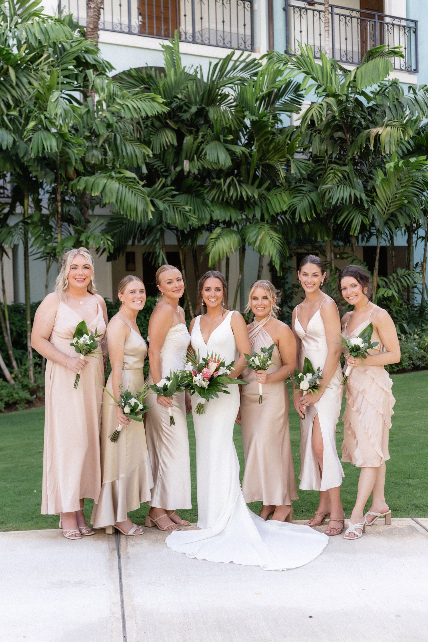 Bridal party poses in champagne-colored dresses holding floral bouquets against tropical palm background.