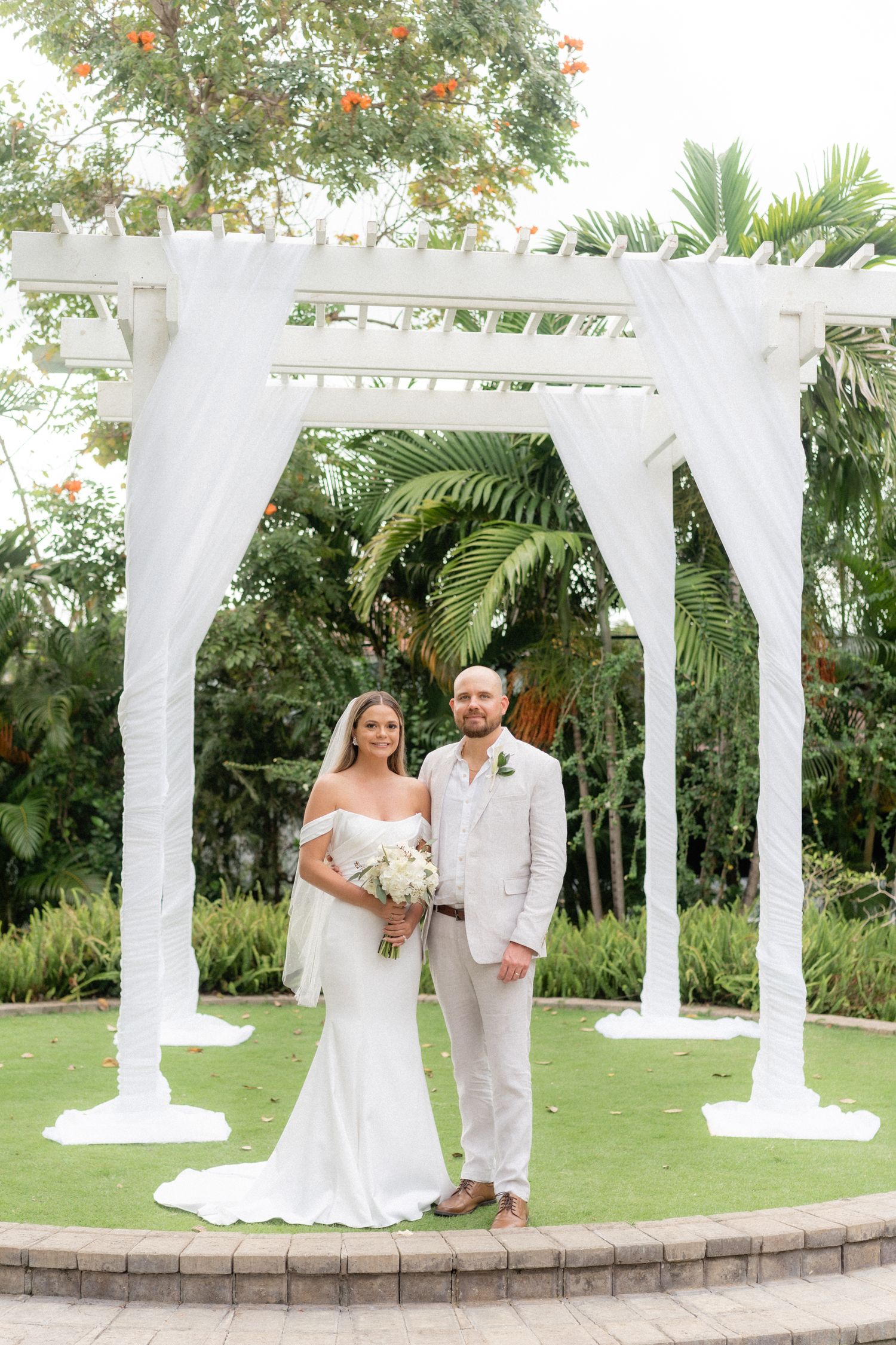 Wedding ceremony on a raised platform with white draped arch and tropical garden backdrop.