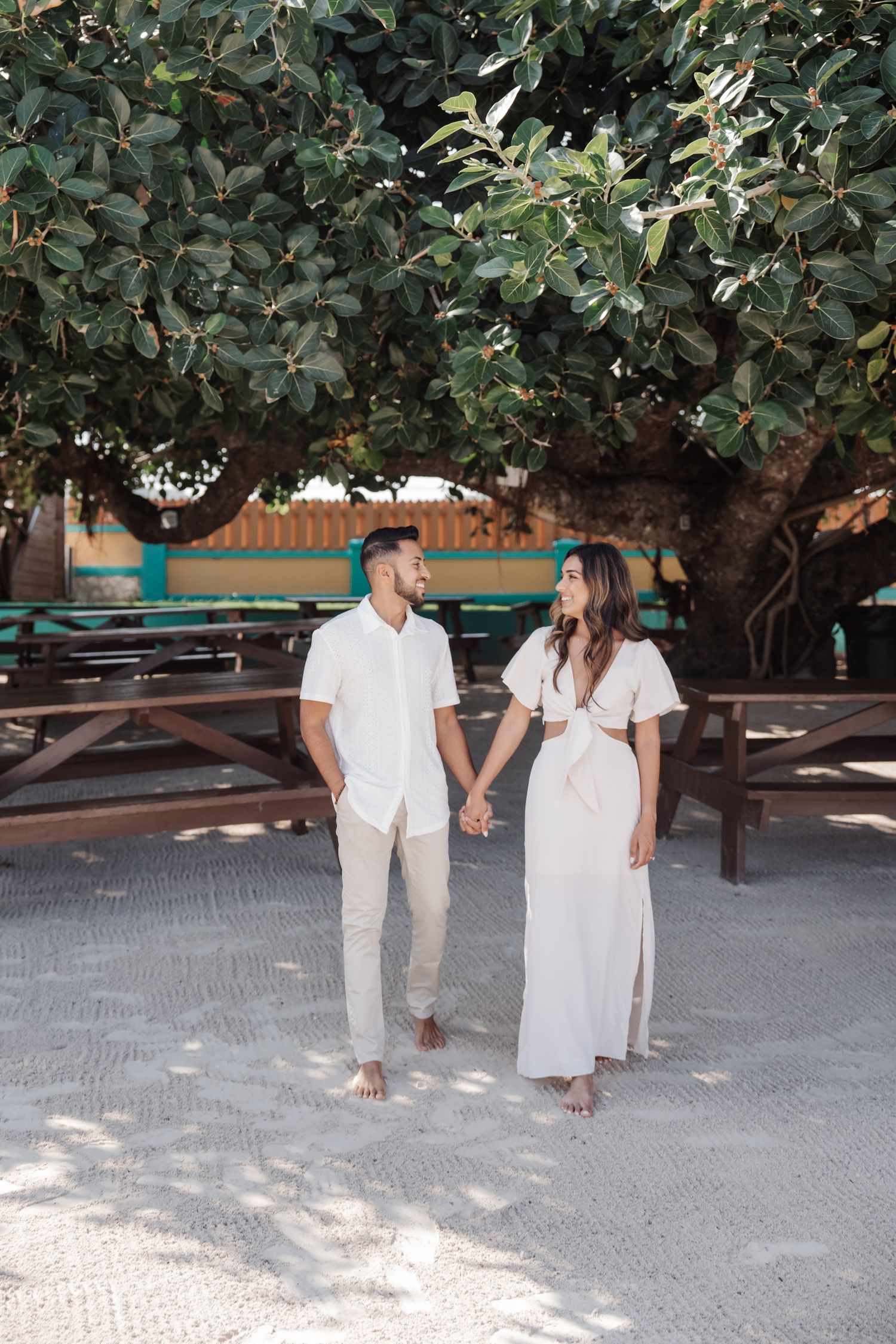 A couple in coordinated white outfits stands beneath shady trees near wooden picnic benches.
