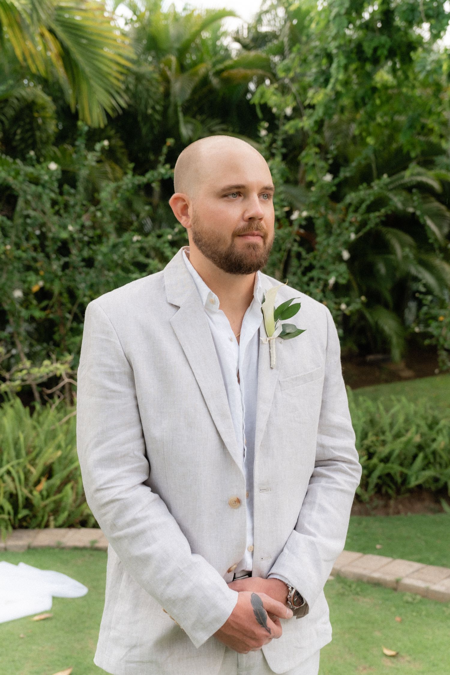 Groom in light gray suit with white boutonniere stands in tropical garden setting.