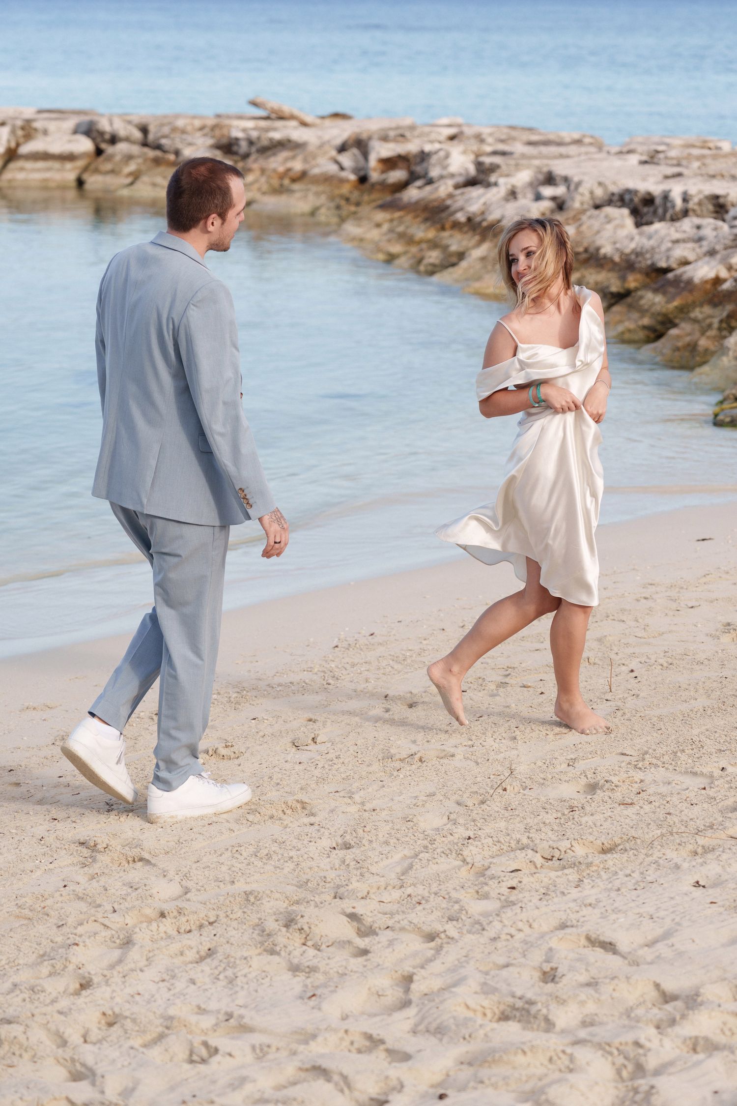 Playful beach wedding moment with couple in grey suit and white dress by rocky shoreline.