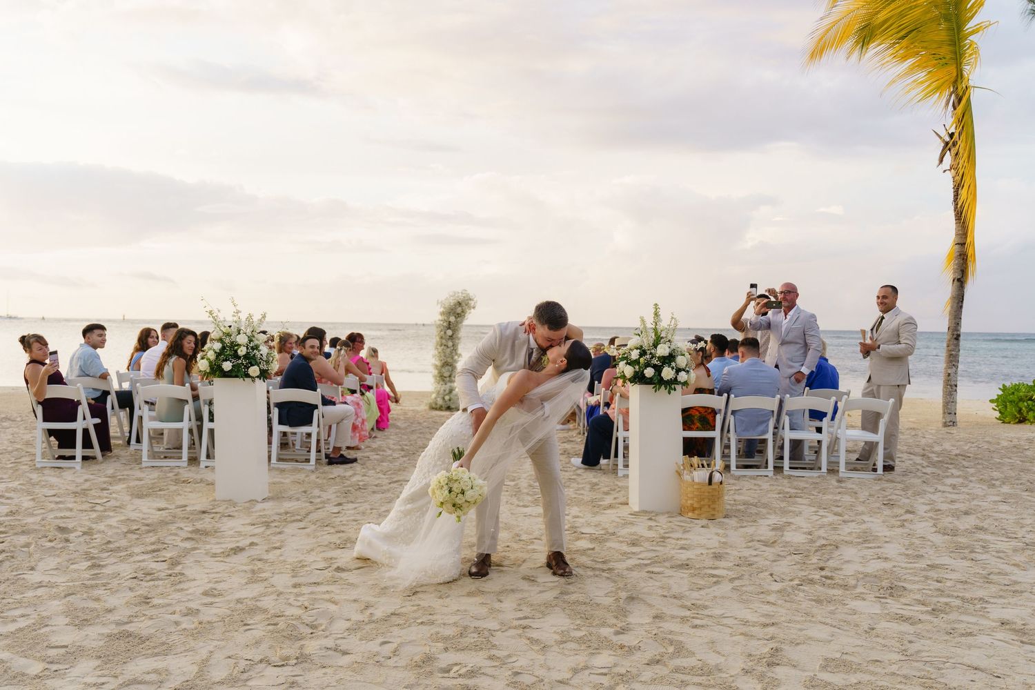 Palm trees sway in the wind at a tropical beach wedding ceremony with white chairs arranged on soft sand.