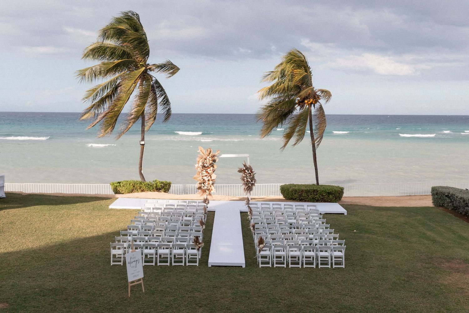 Oceanfront wedding ceremony setup with white chairs and palm trees swaying in the wind.