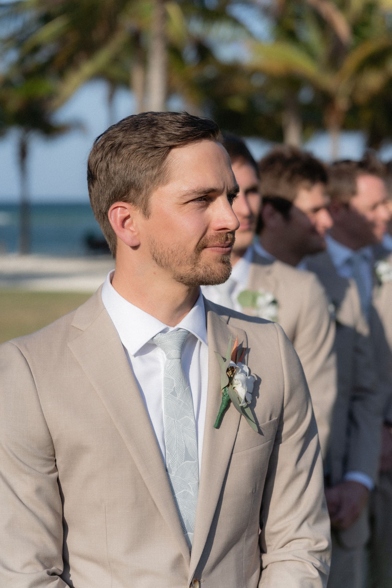 A groomsman in a beige suit with boutonniere stands during outdoor beach ceremony.
