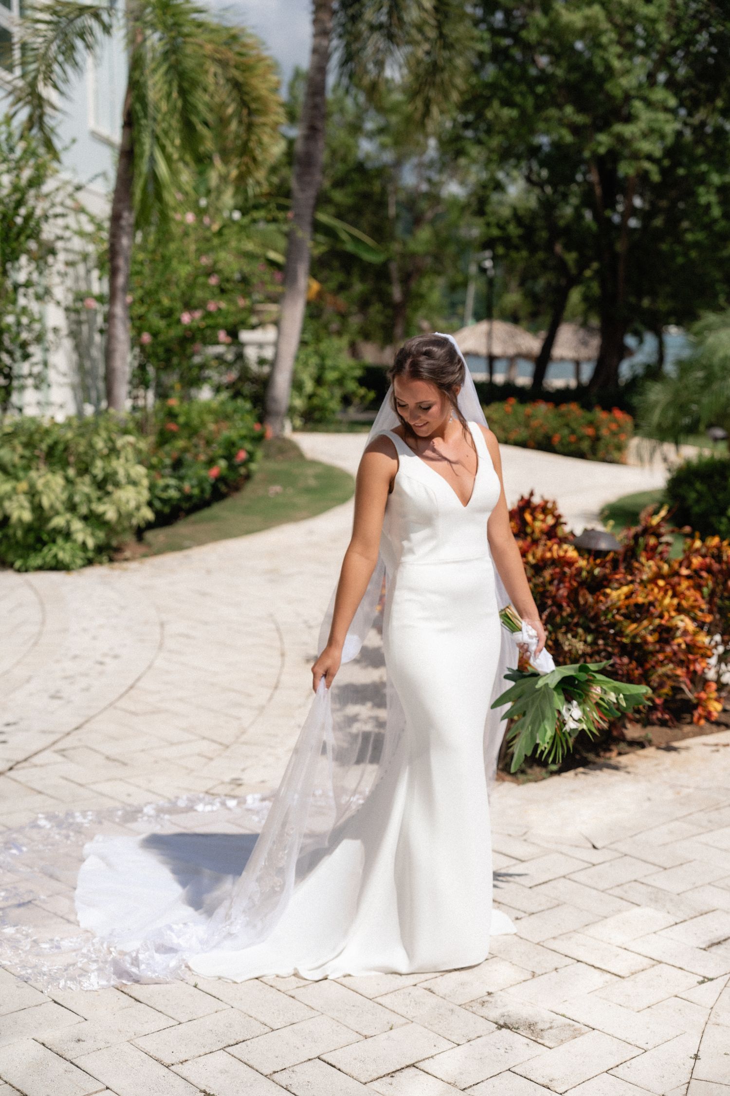 Elegant bride in fitted white wedding dress poses on stone path surrounded by tropical garden landscaping.