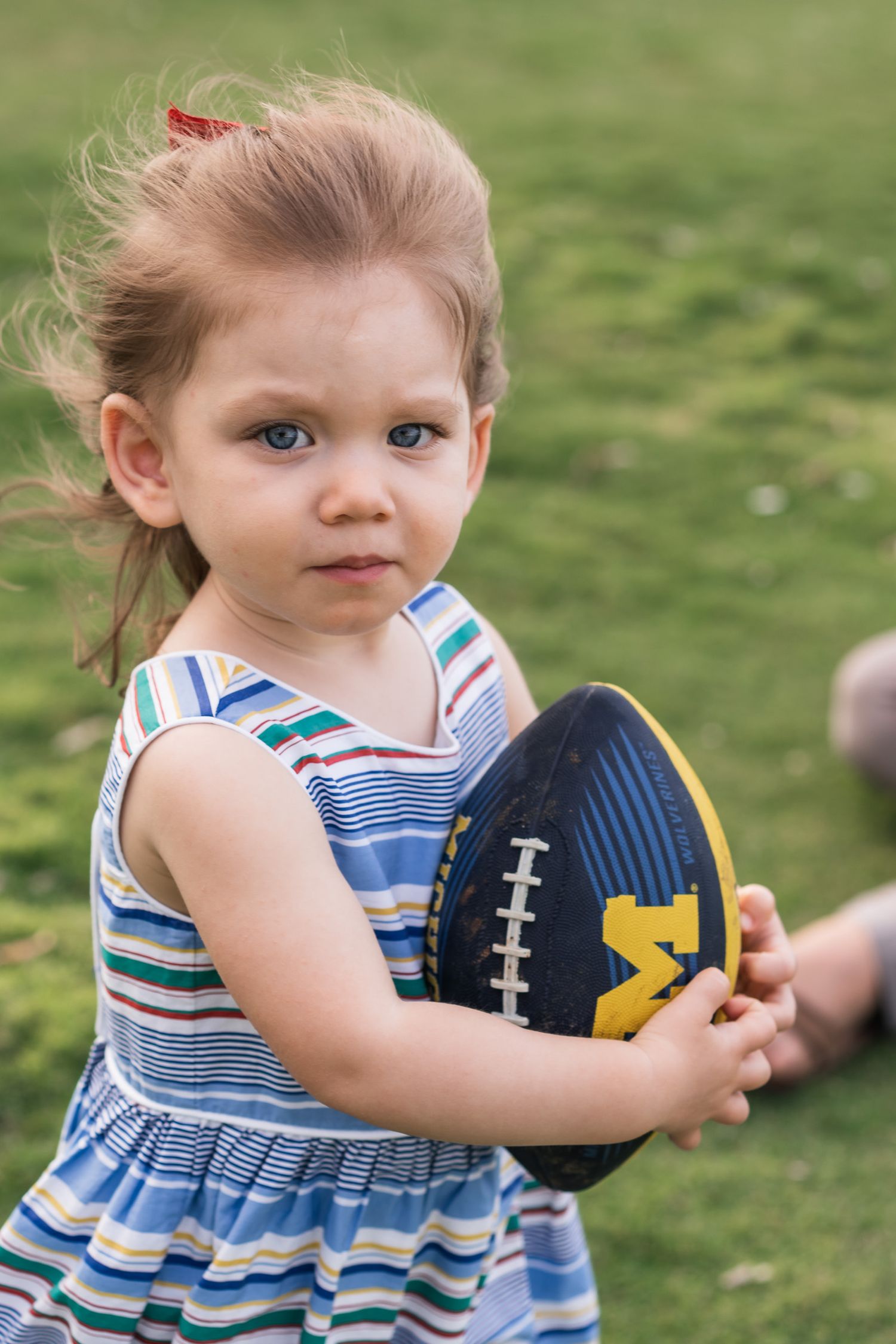 Young child in striped dress holding a blue and yellow football on grass.