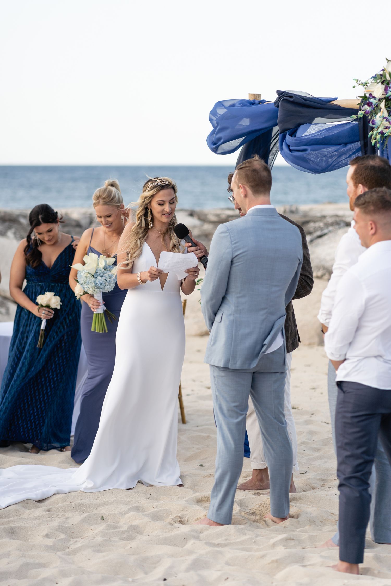 Beach wedding ceremony with bride reading vows under navy blue draped arch.