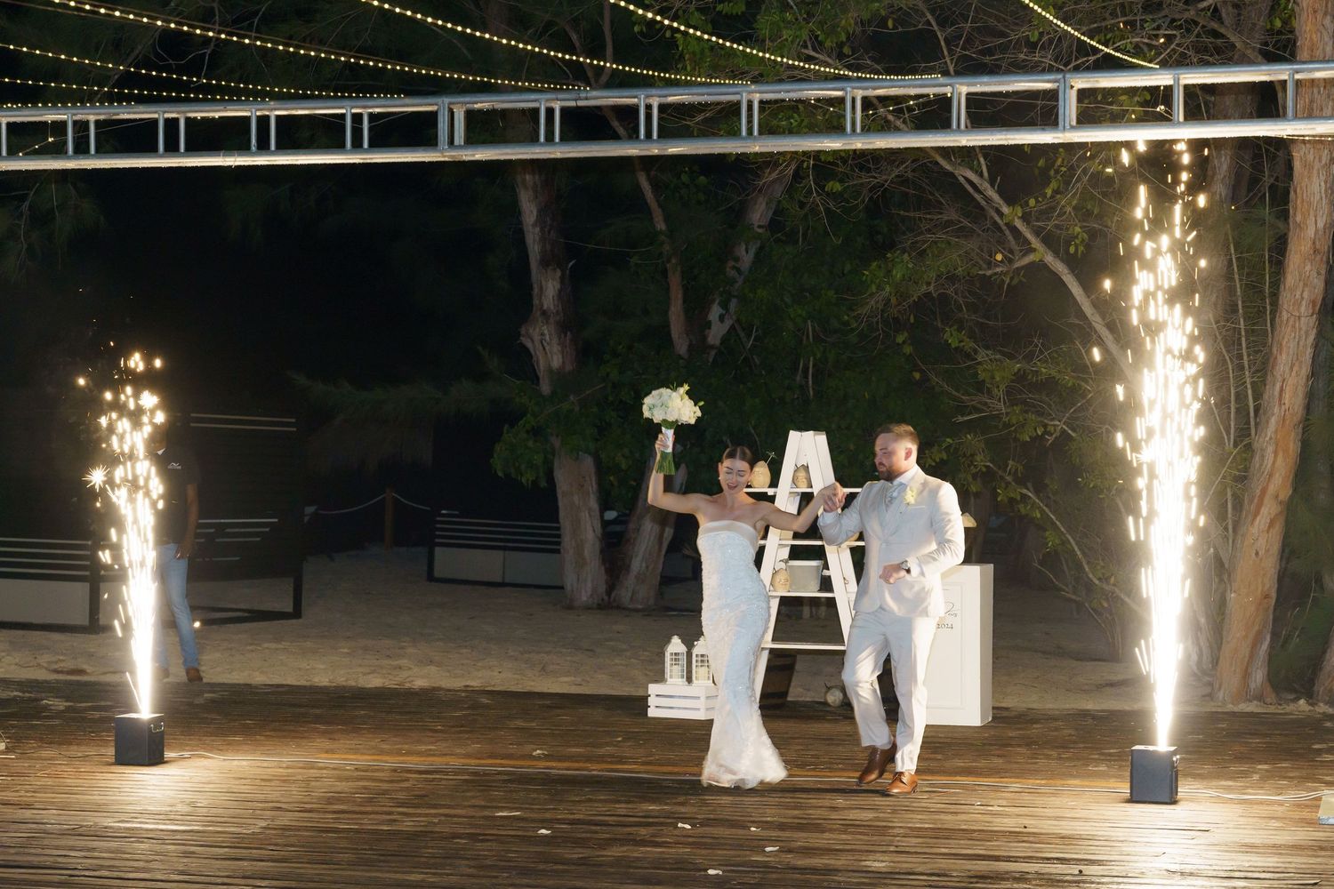 Sparklers illuminate an elegant nighttime beachside wedding ceremony setup with white decor on a wooden deck.