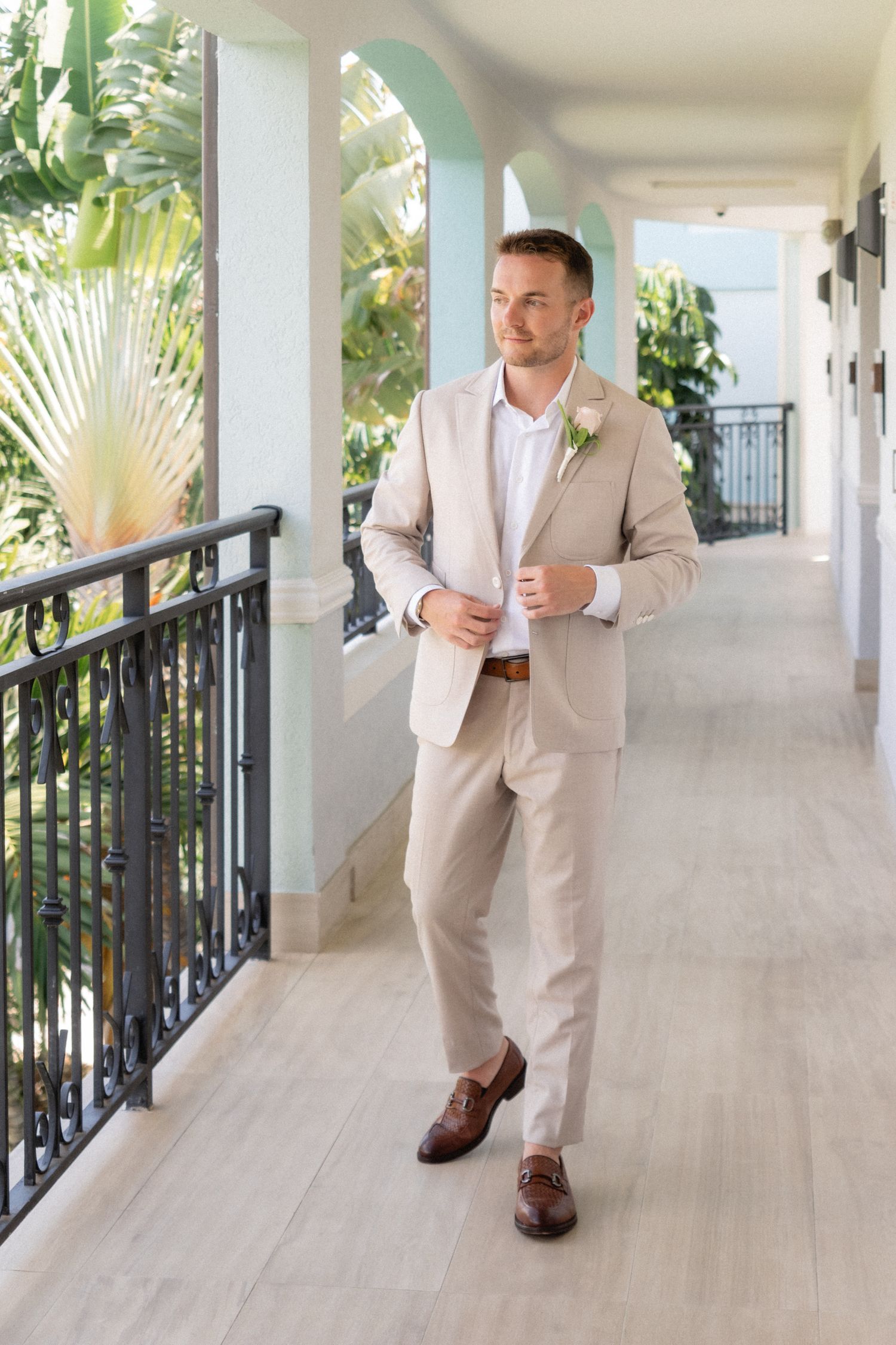 Groom in light beige suit walking along a covered resort balcony with tropical landscaping.