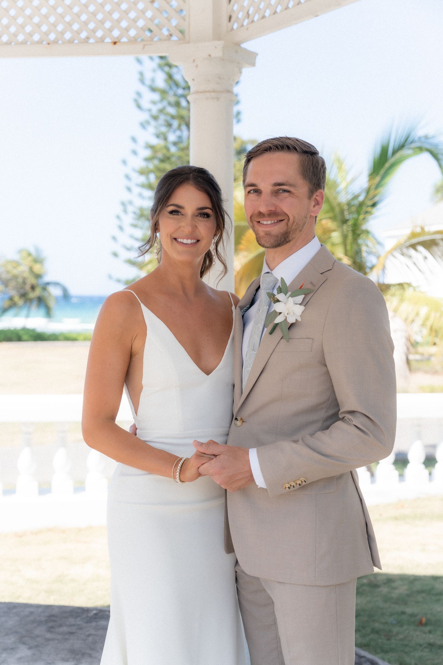 A couple in wedding attire poses under a gazebo with palm trees and ocean views in the background.