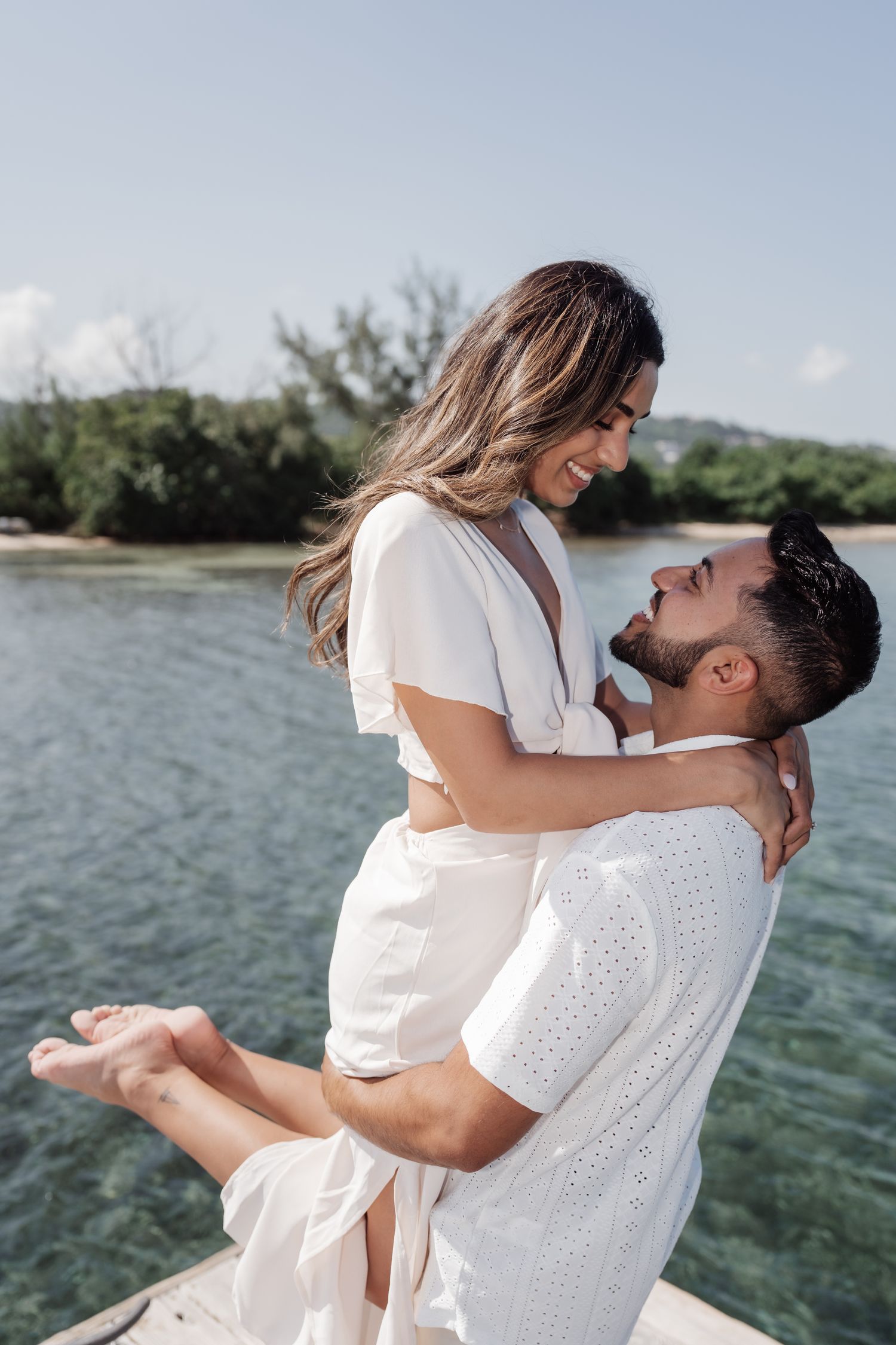 Couple in white outfits sharing an intimate moment by the waterfront with lush greenery in background.