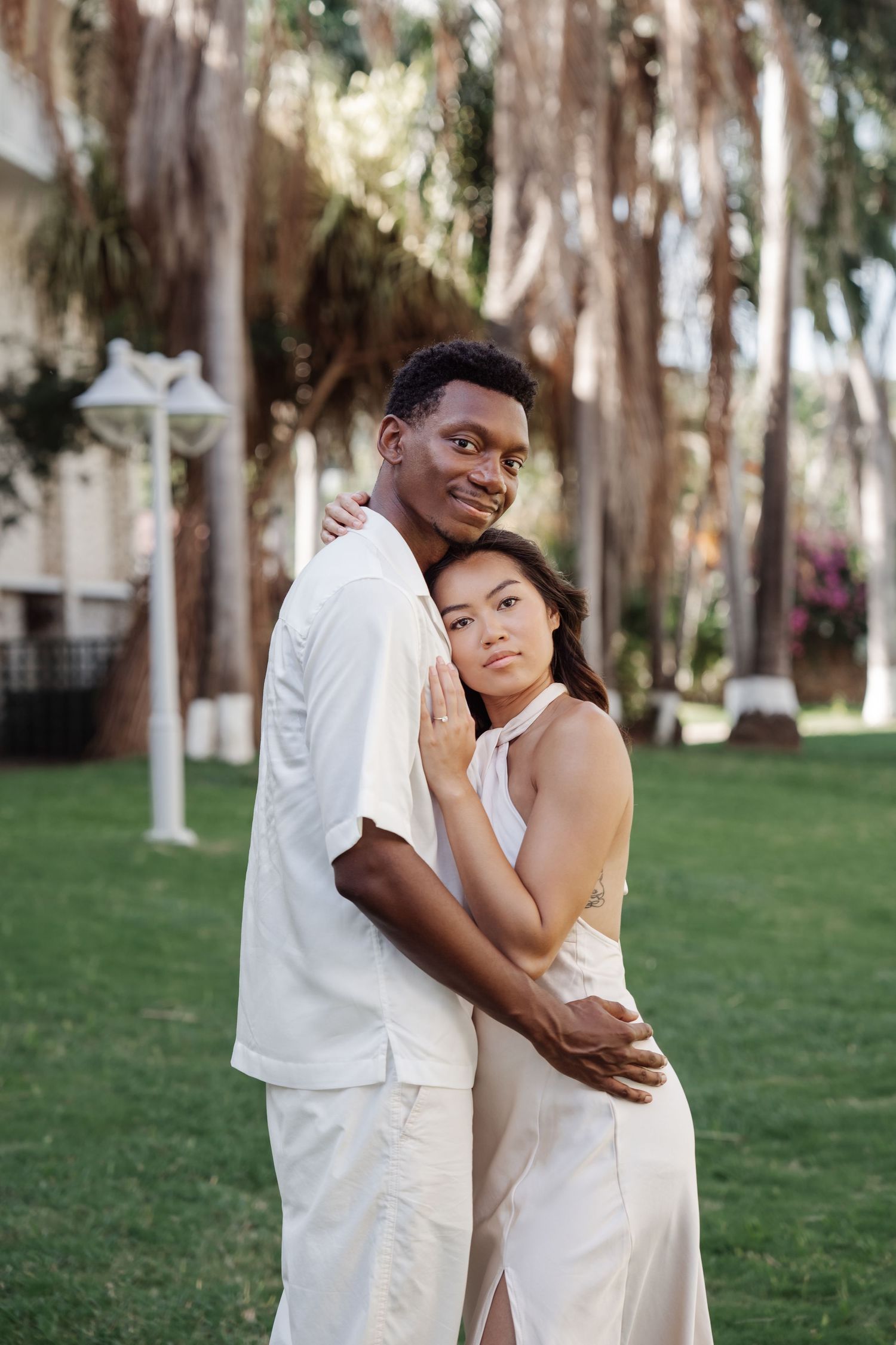 A romantic couple in white outfits embraces on a lush garden path lined with tall palm trees.