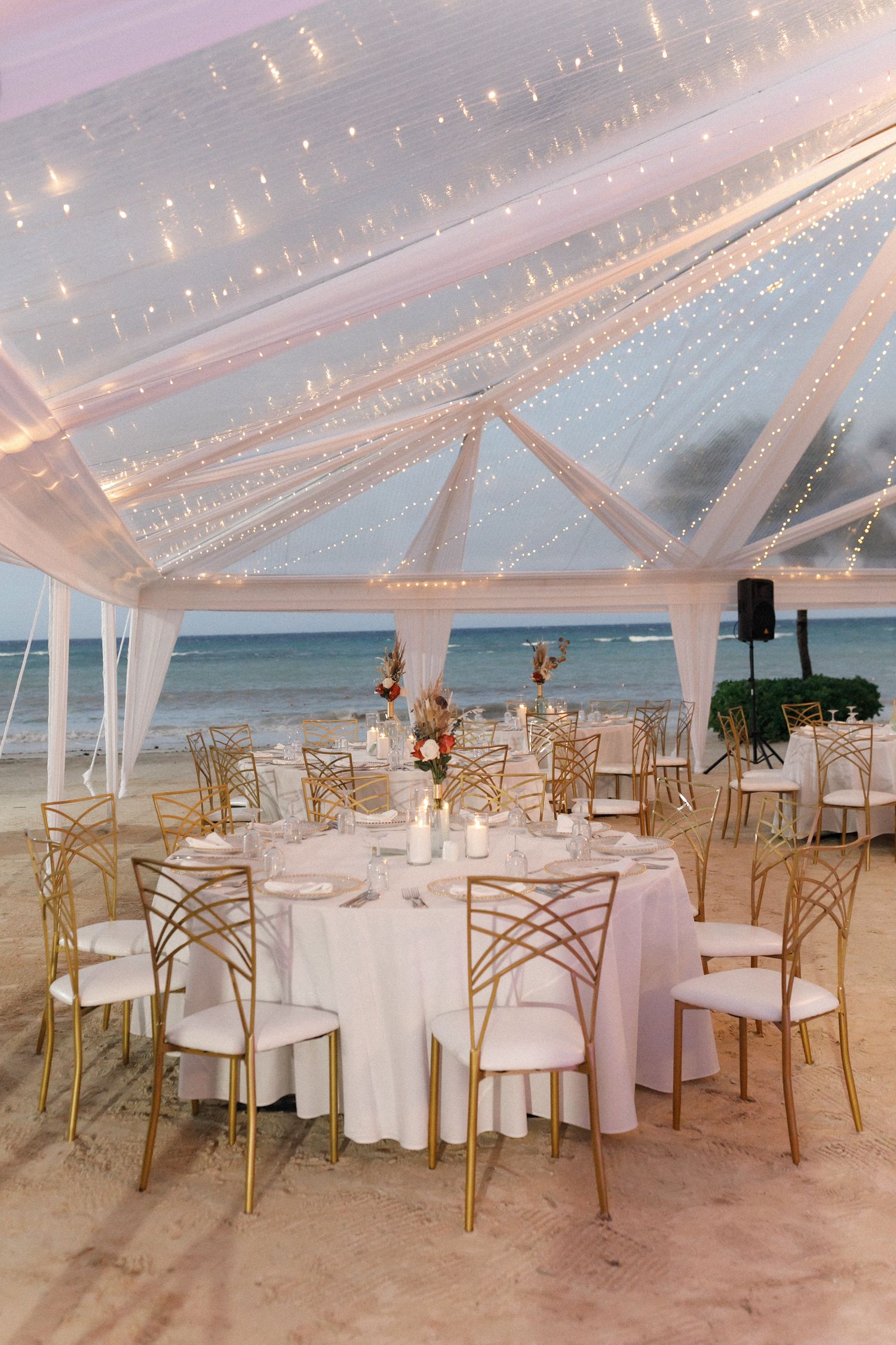 Elegant beach wedding reception setup with white tent, string lights and gold chairs overlooking the ocean at sunset.