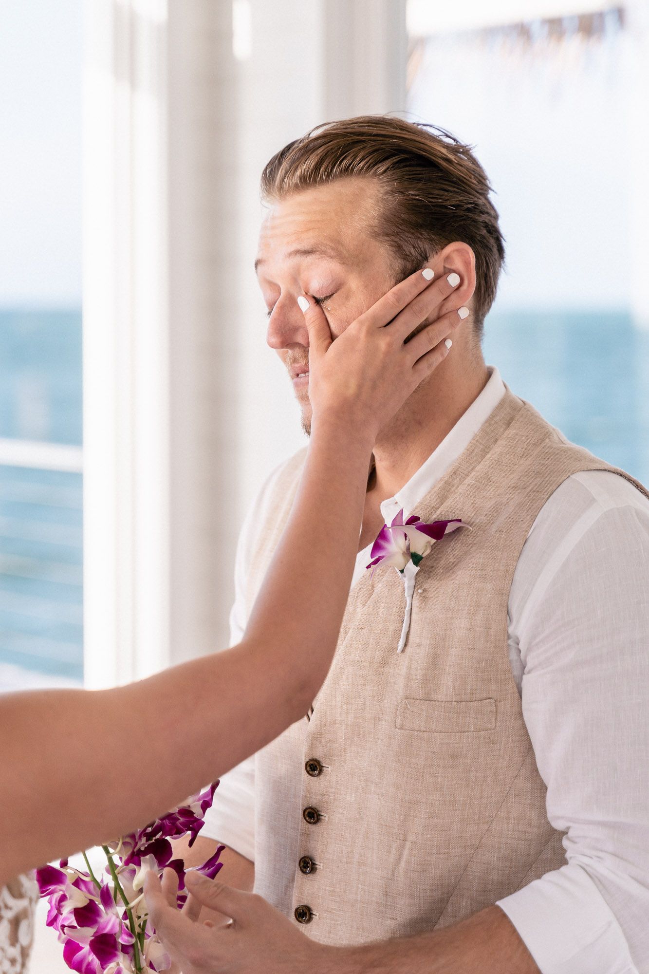 An emotional wedding moment with a groom in a beige vest wiping his eyes.