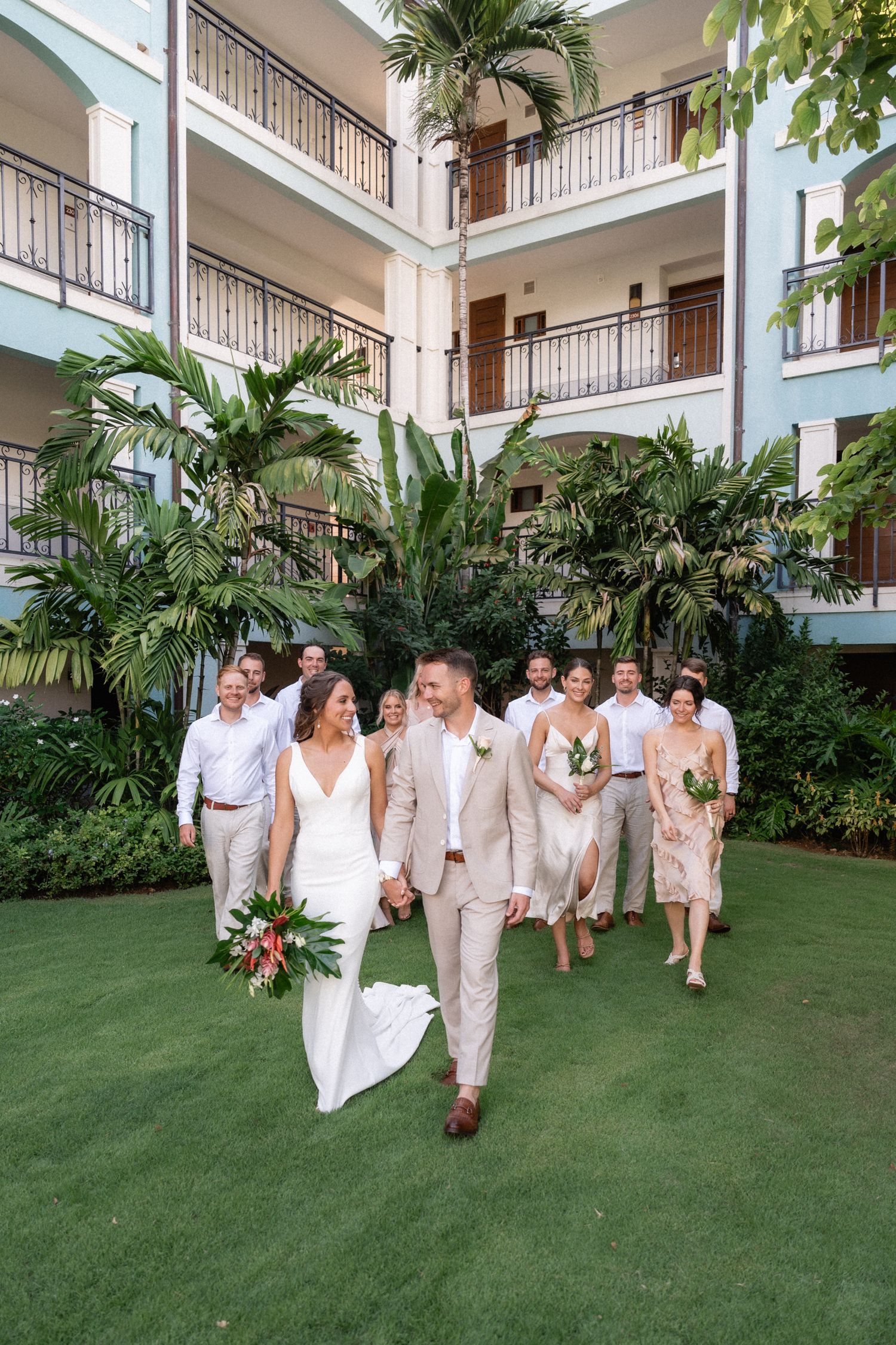 Wedding party walking on grass courtyard surrounded by tropical resort architecture.