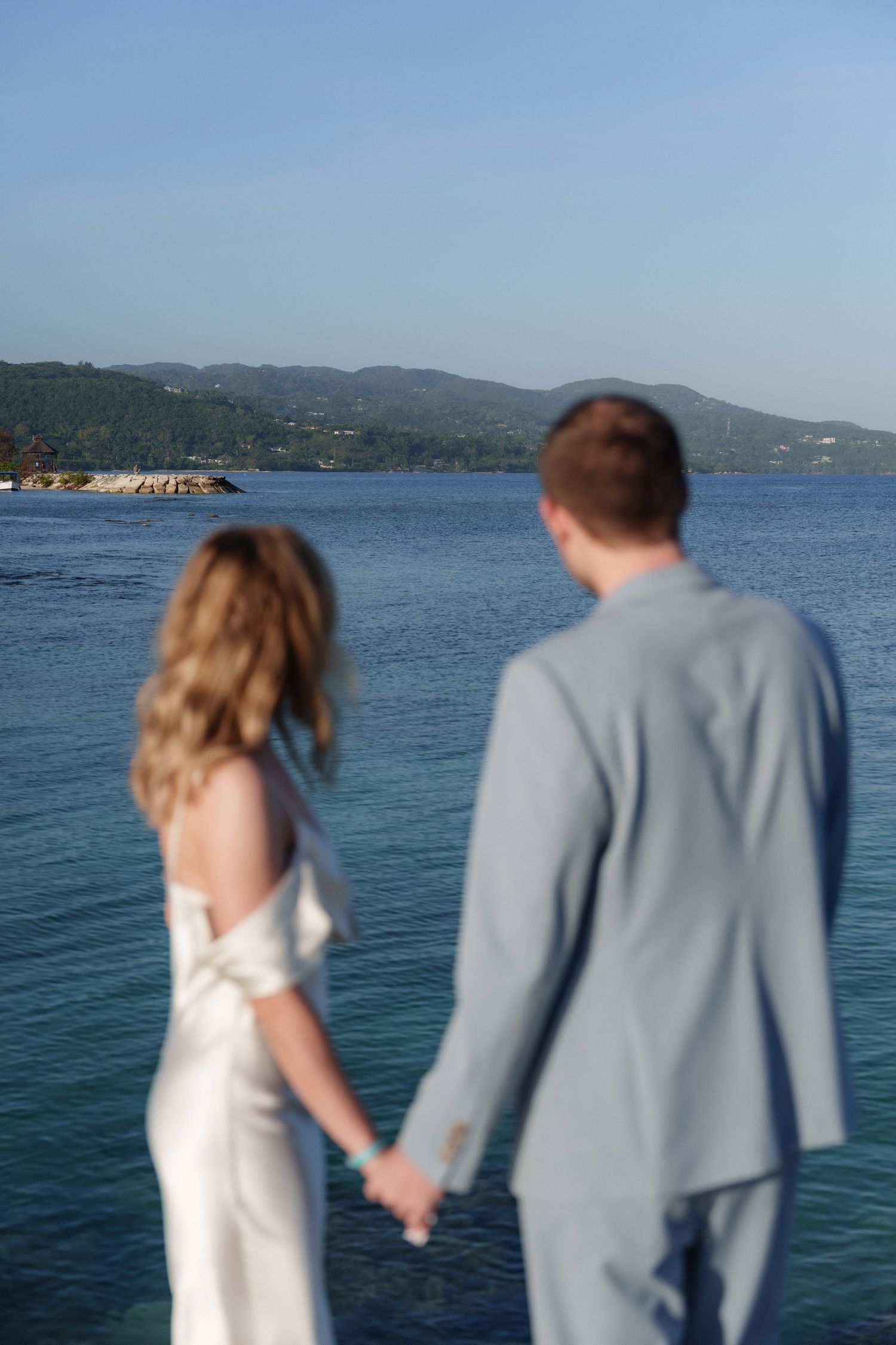 A couple holding hands while gazing at a scenic ocean view with mountains in the background.