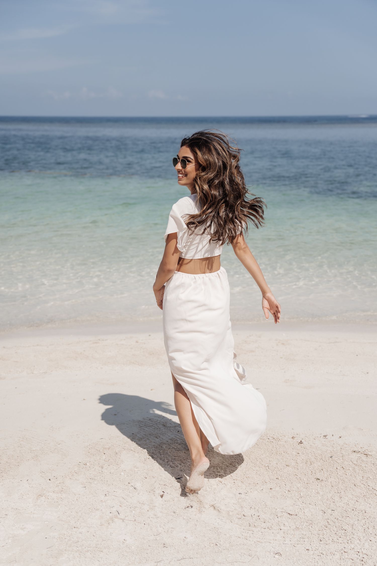 Woman in a flowing white dress walking on a tropical beach with windswept hair.