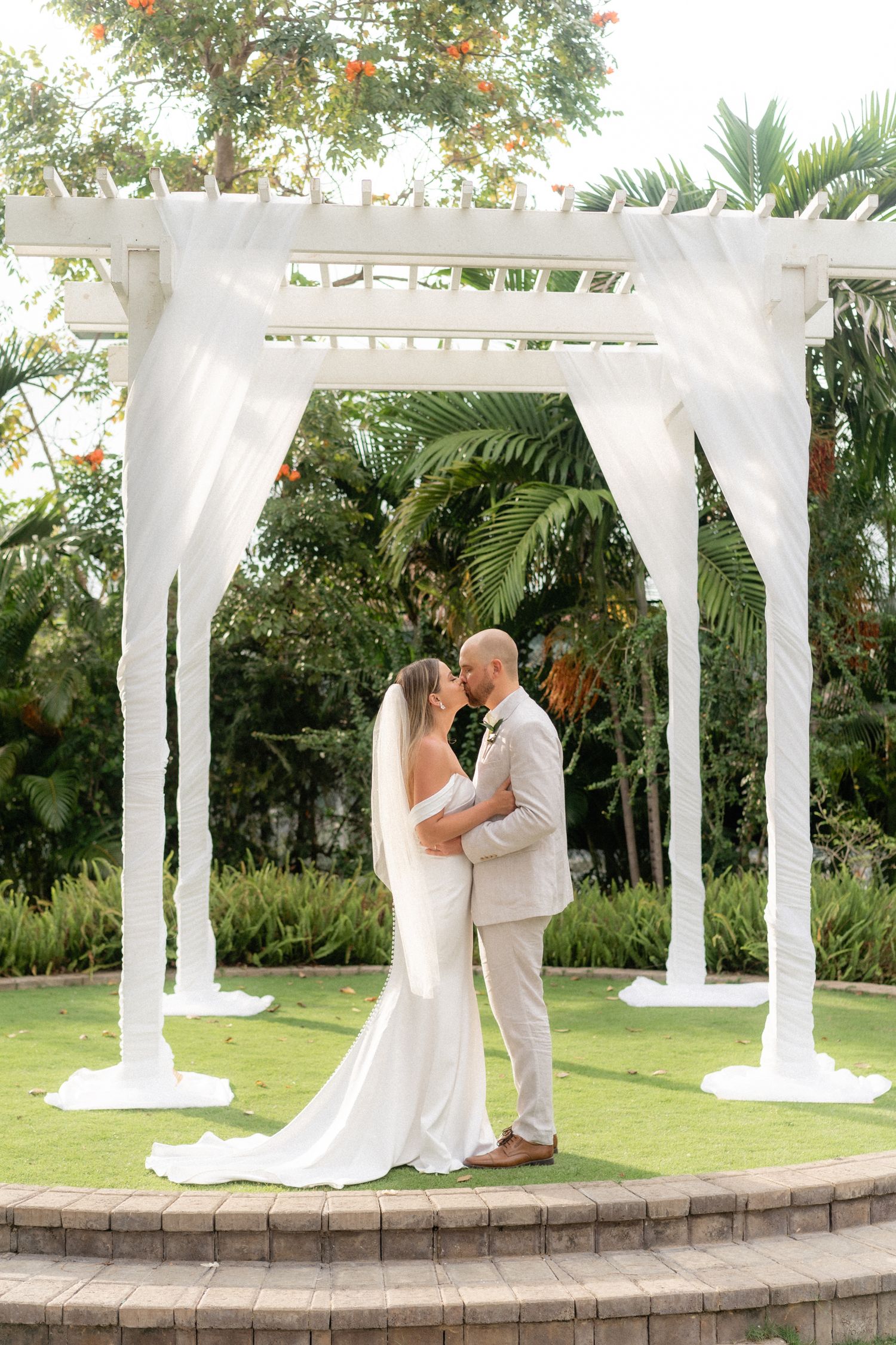 A romantic wedding ceremony under a white draped pergola surrounded by lush tropical gardens.