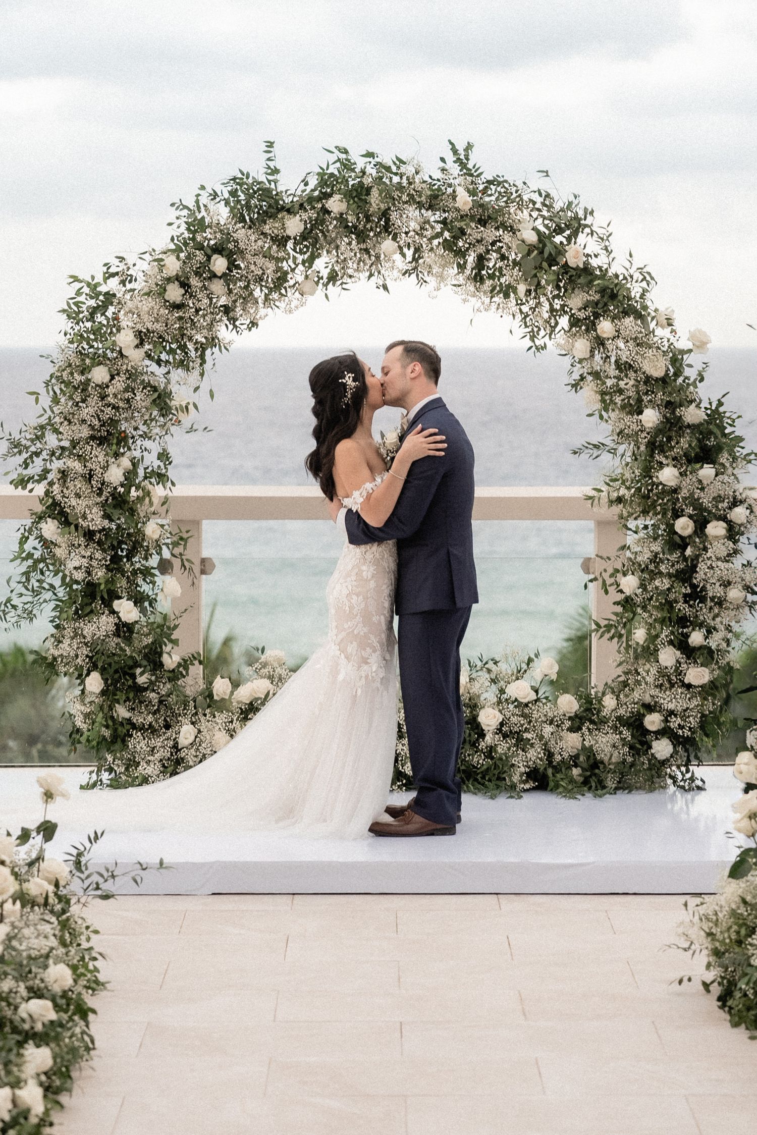 A romantic wedding kiss under an ornate floral arch against an ocean backdrop.