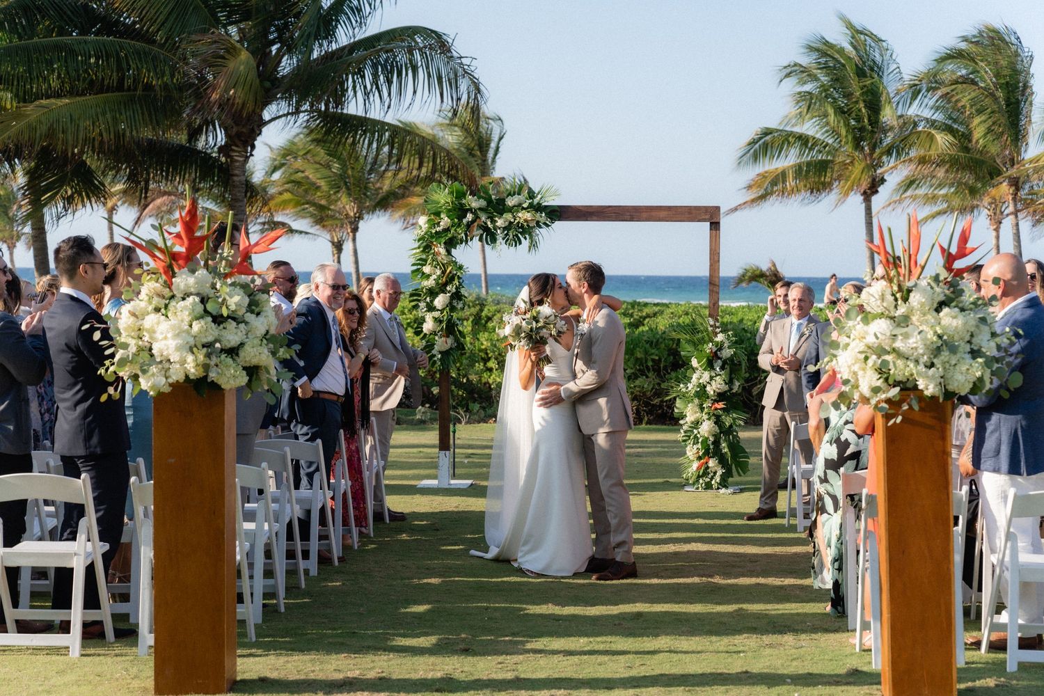 Wedding ceremony on a tropical lawn with floral arrangements and white chairs under swaying palm trees.