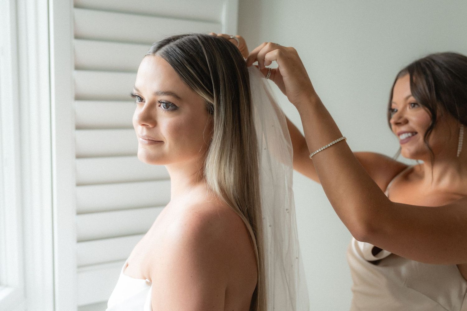 Getting ready scene of bride having her veil adjusted near a window.