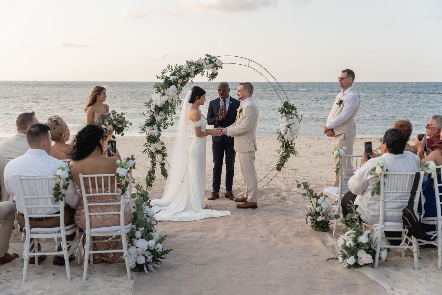 Beachside wedding ceremony with floral arch and white chairs on sandy shore at sunset.