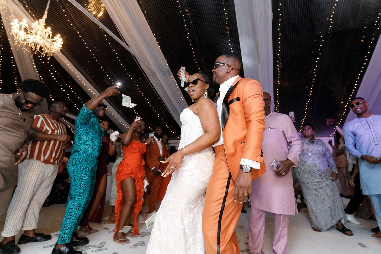 Wedding couple dances surrounded by guests under twinkling lights in a decorated reception tent.