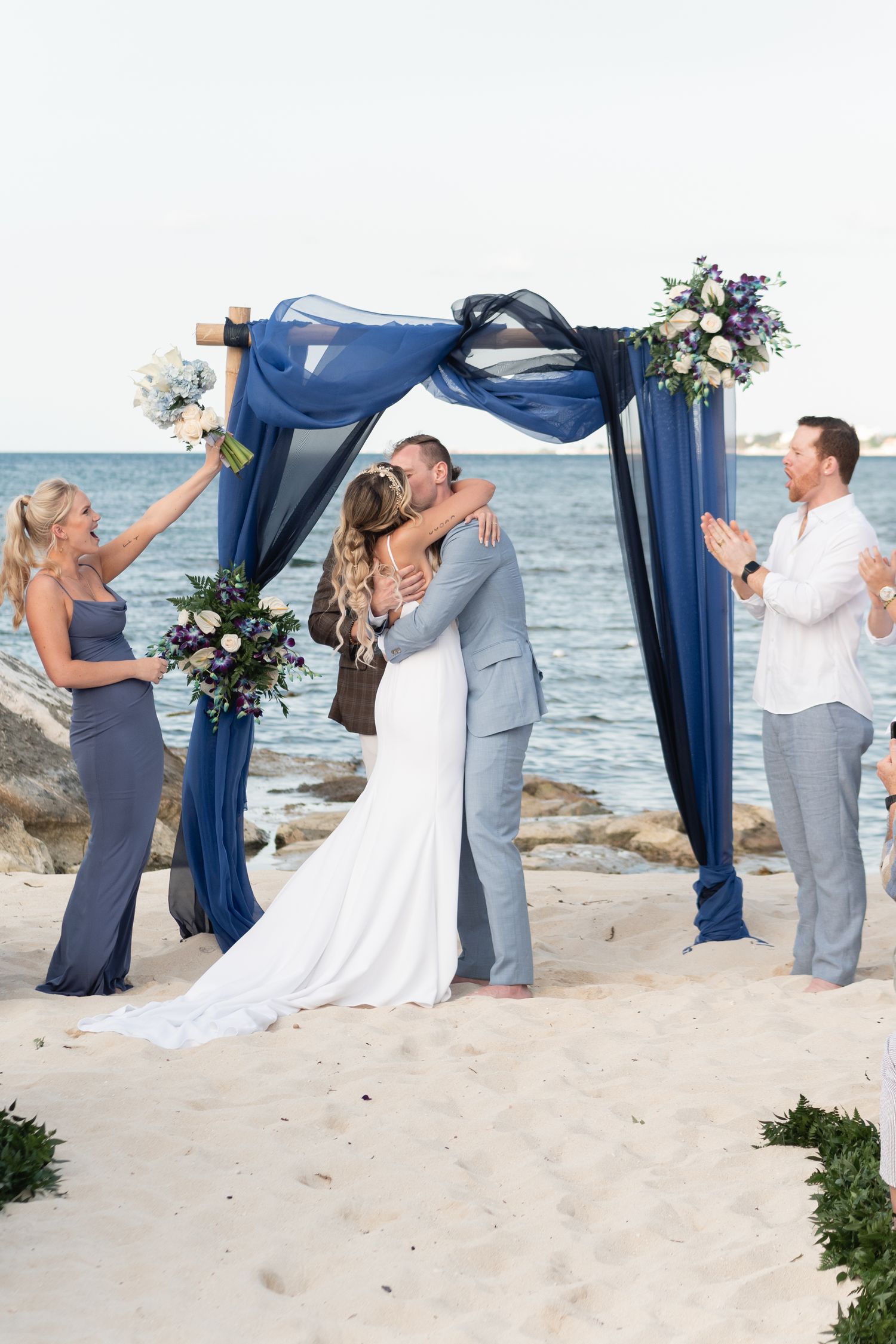 Wedding kiss at beachside ceremony under blue fabric arch with guests celebrating.