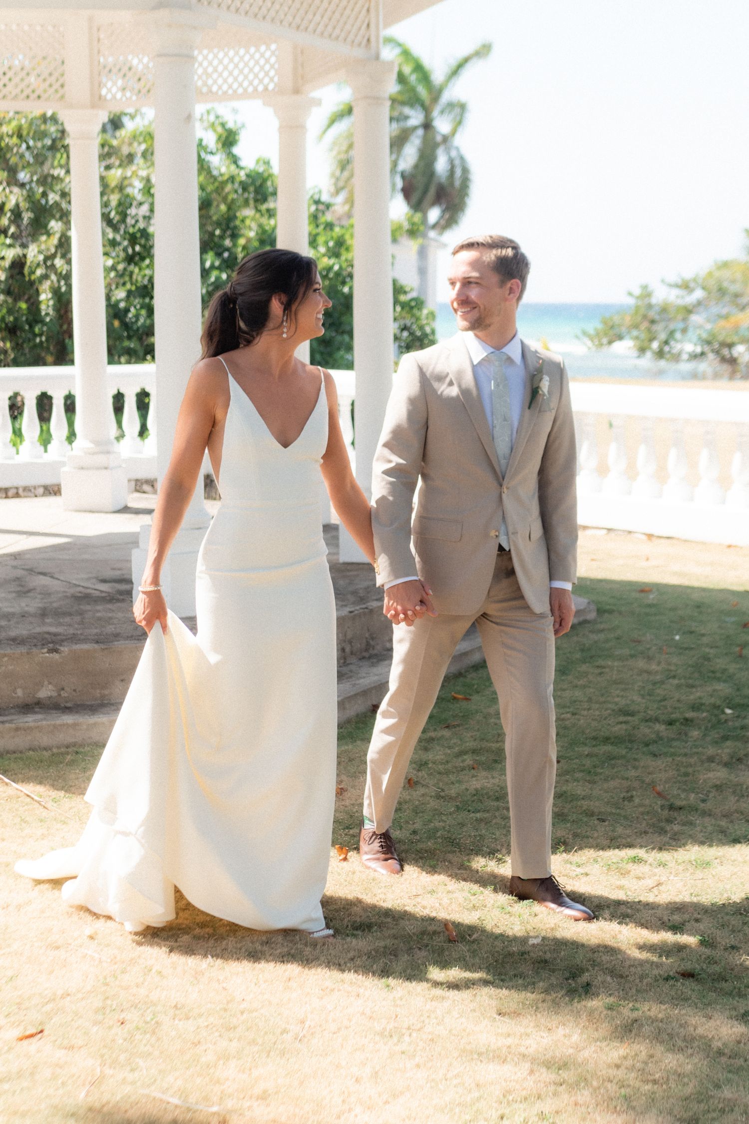 Elegant couple in wedding attire holding hands under white gazebo with ocean backdrop.