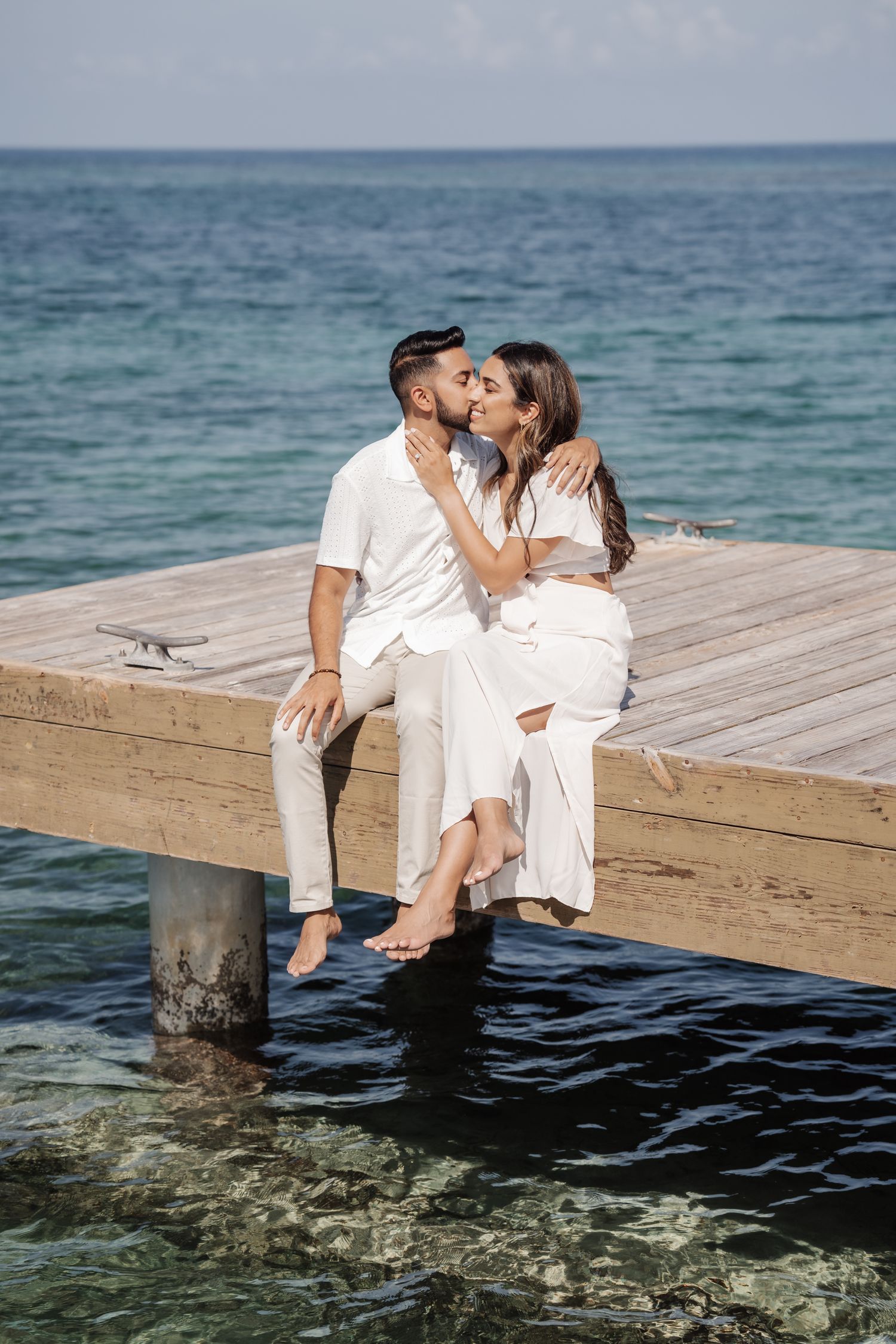 Couple in white outfits sitting romantically on a wooden pier over crystal clear waters.