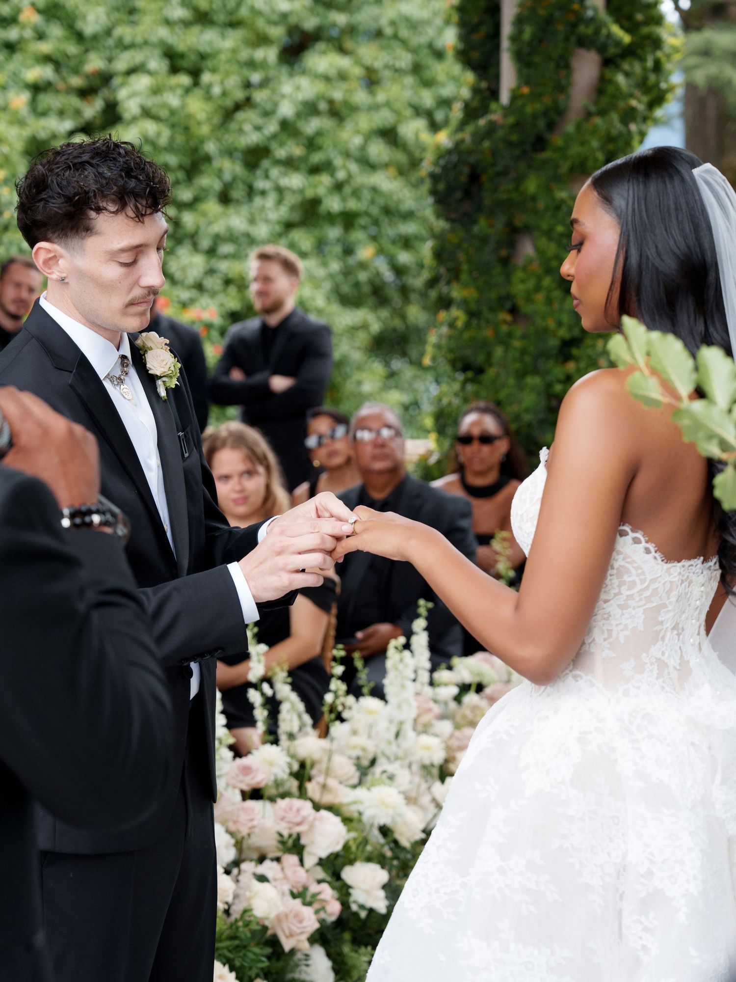 A couple exchanges rings during an outdoor wedding ceremony surrounded by white floral arrangements and guests.