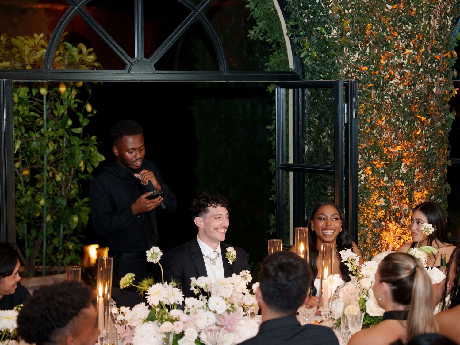 A wedding reception dinner table setting with elegant white floral centerpieces and glowing candles under glass windows.