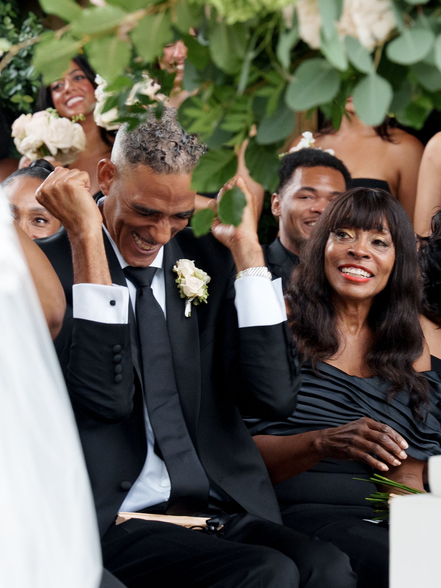 Wedding guests sharing joyful laughter during outdoor celebration with greenery in background.