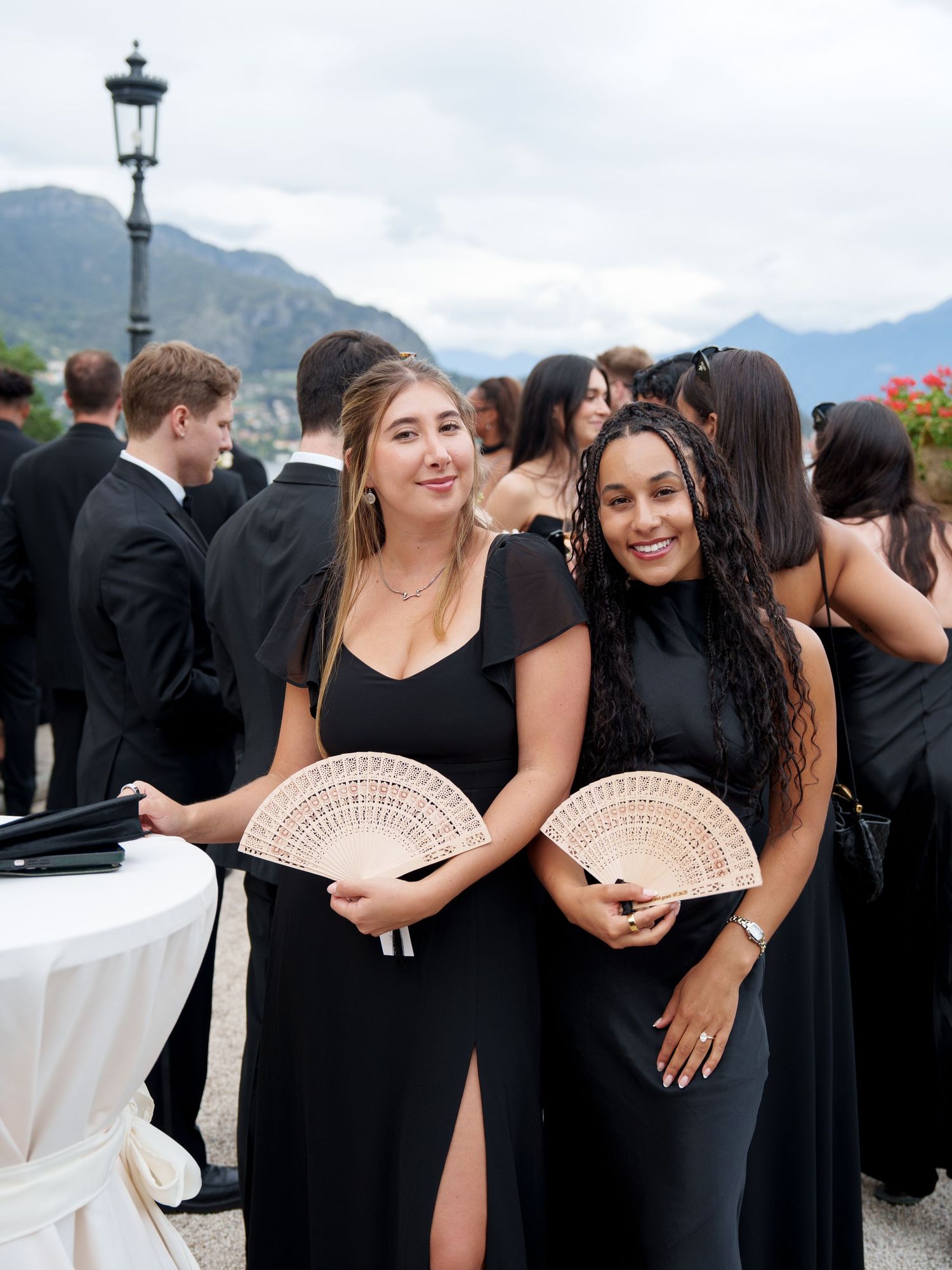 Bridesmaids in elegant black dresses pose with decorative fans at a lakeside wedding with mountains in the background.
