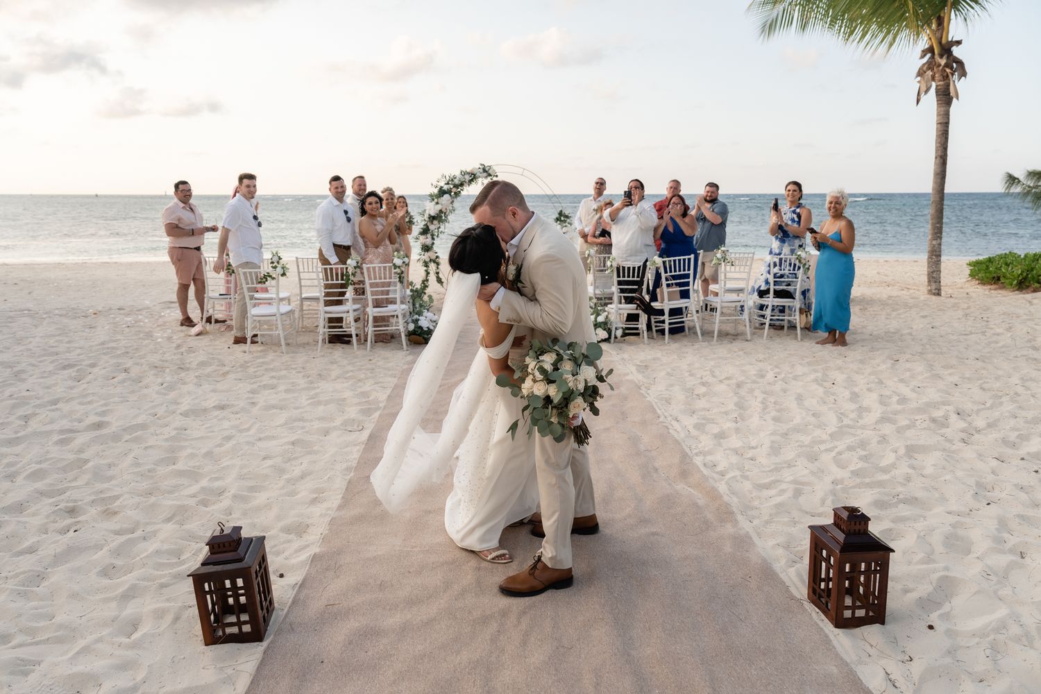 A seaside wedding ceremony on the beach with guests seated and decorative lanterns in the sand.