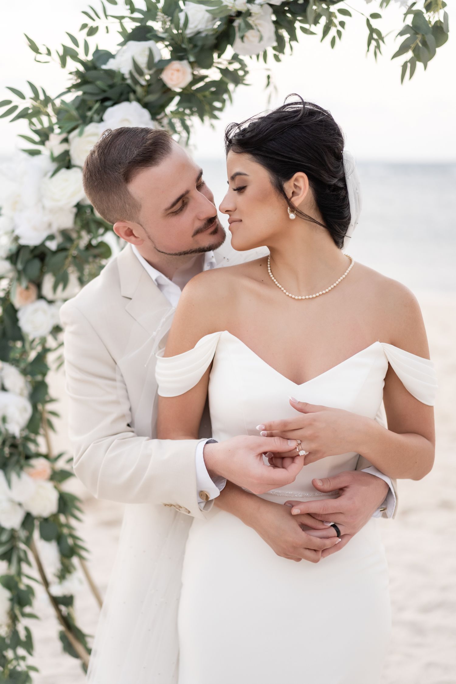 Wedding couple poses under floral arch with white roses and greenery on beach.