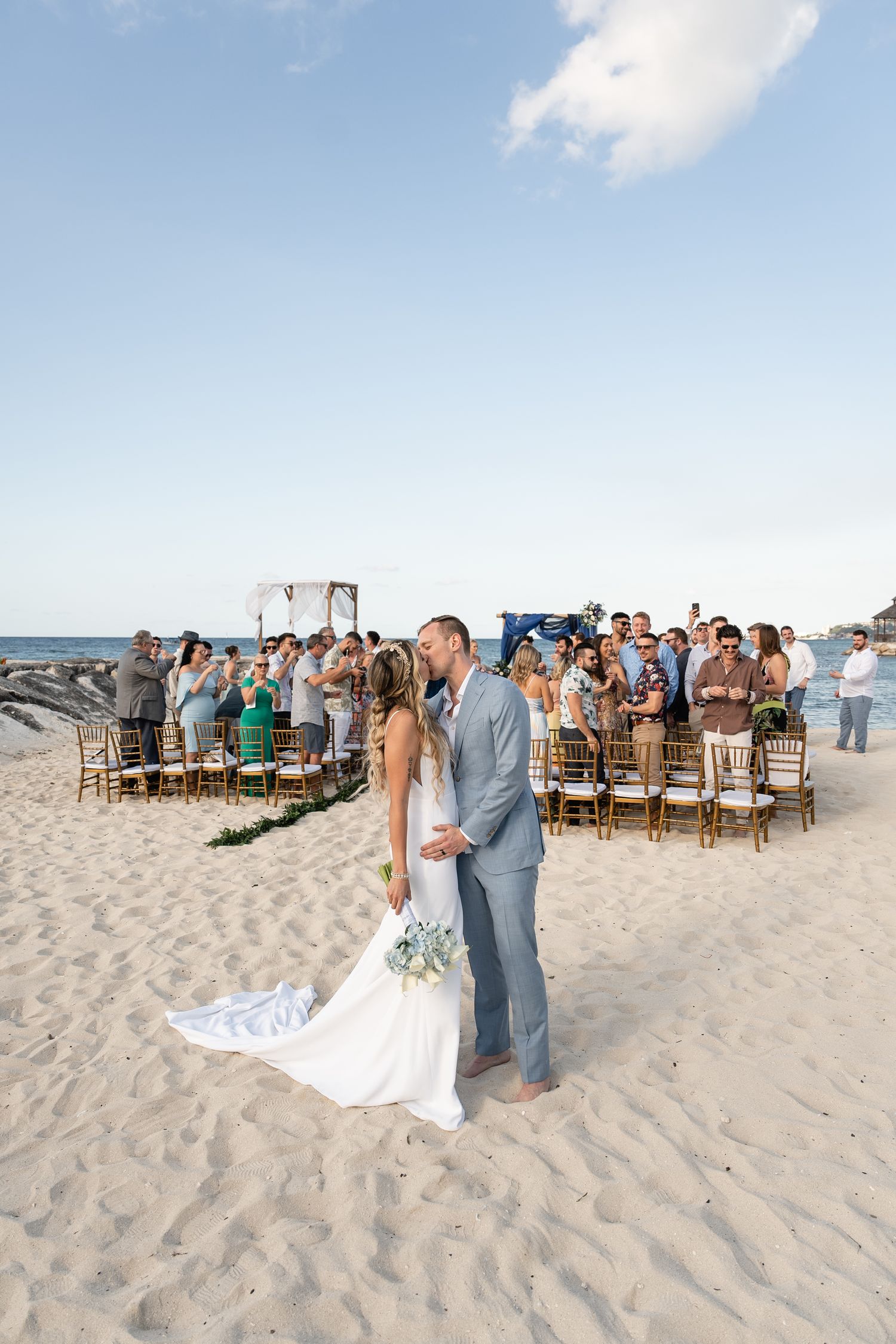 Elegant beach wedding ceremony with guests seated on wooden chairs watching couple against ocean backdrop.