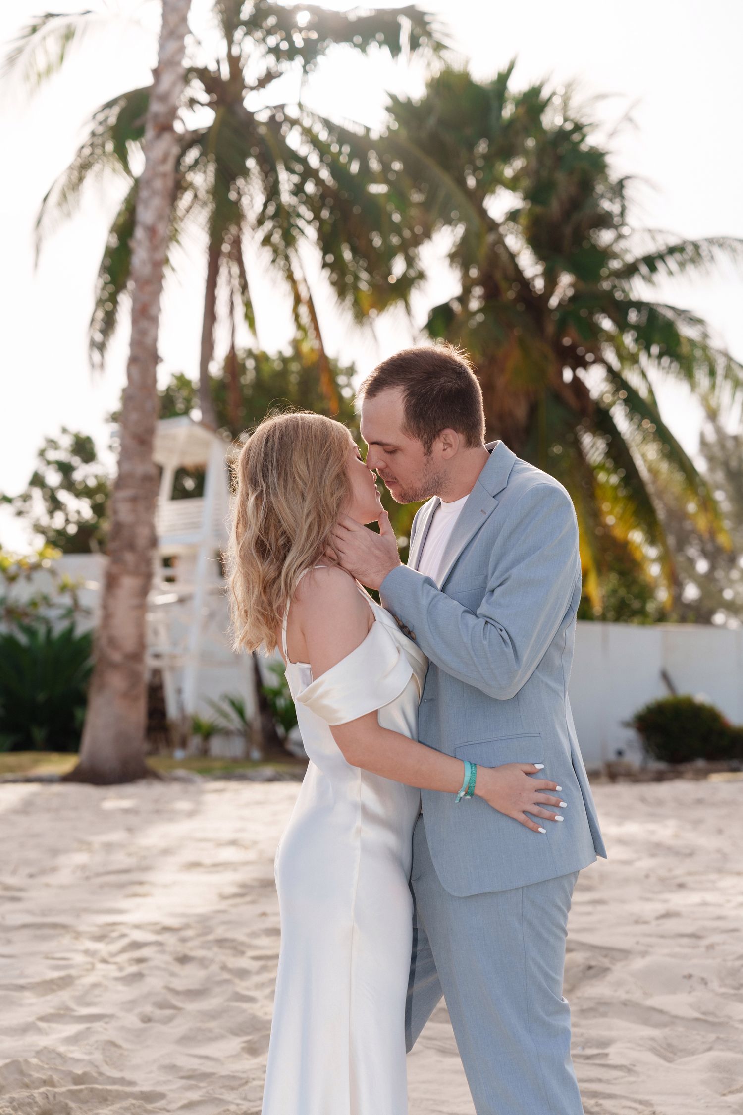 Couple embracing on beach with palm trees and white architecture in background.
