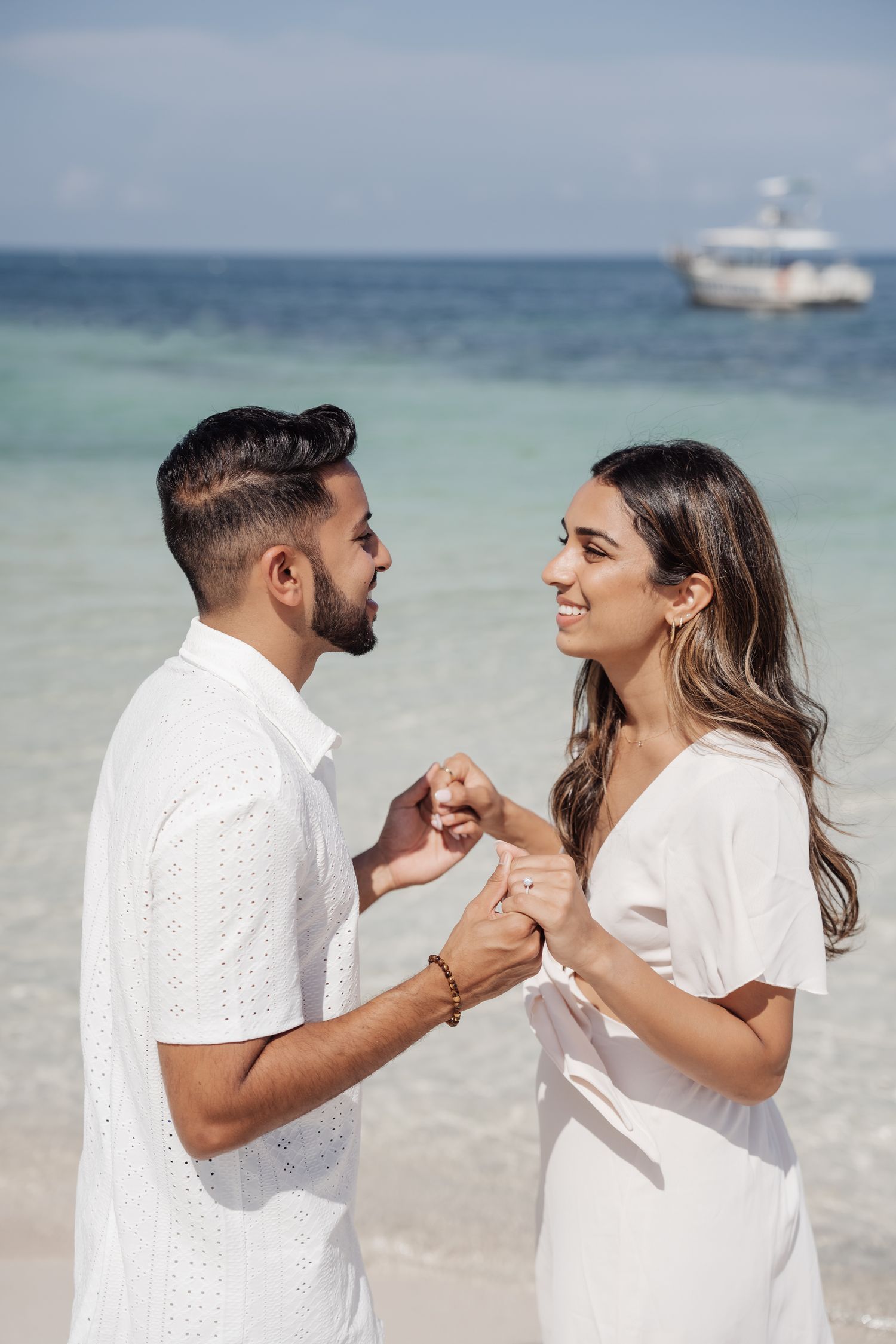 A romantic couple shares an intimate moment on a tropical beach with a boat visible in the distance.
