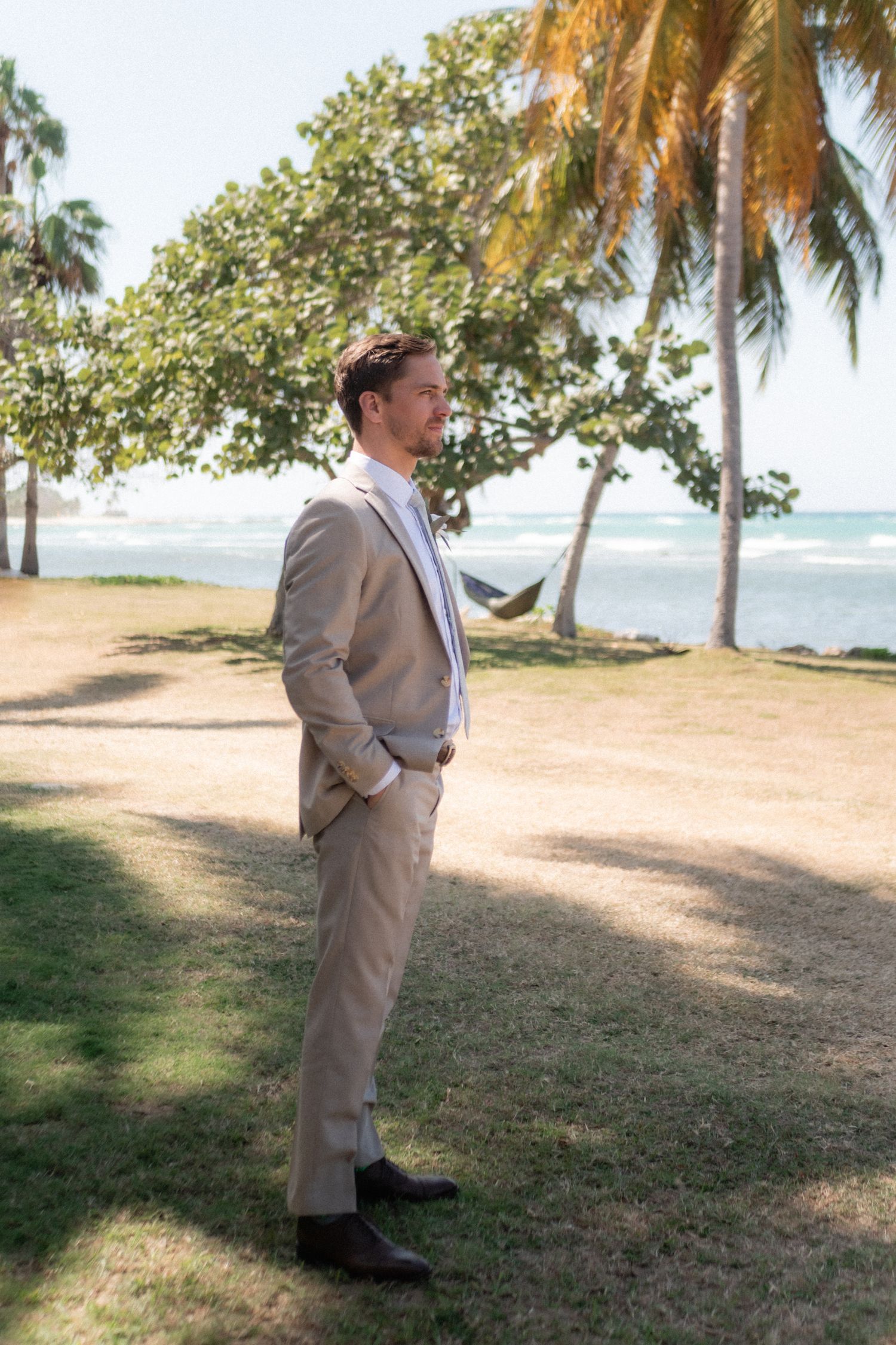 Groom in beige suit stands contemplatively near palm trees and ocean view.
