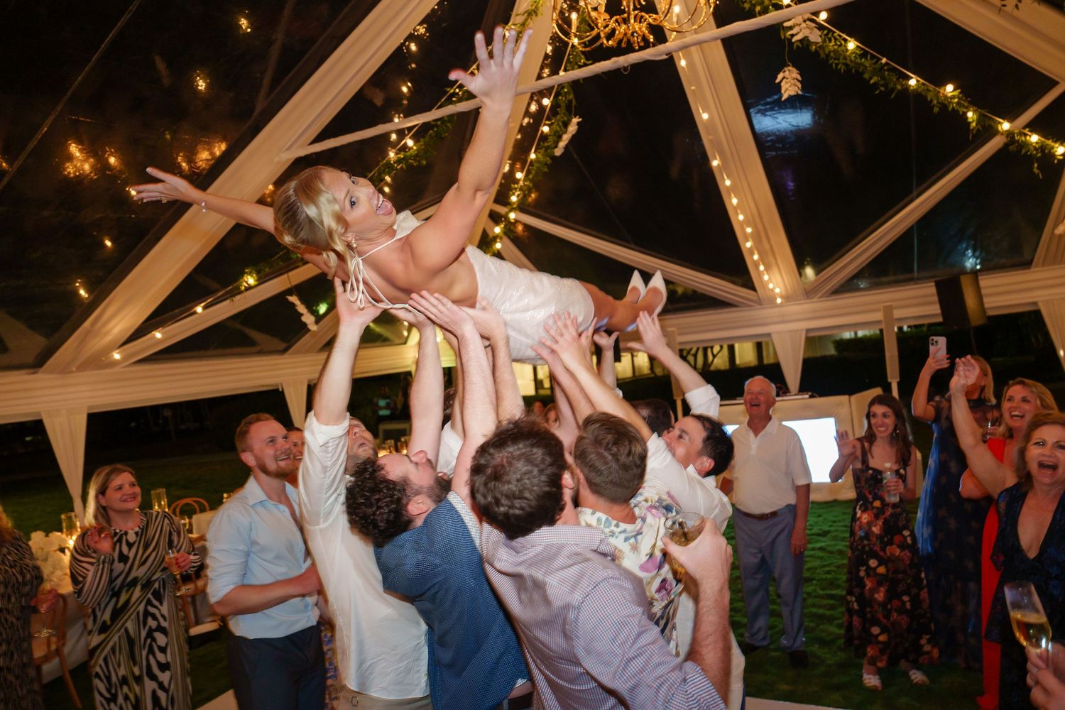 Wedding celebration with guests lifting someone in the air under a decorated tent ceiling.