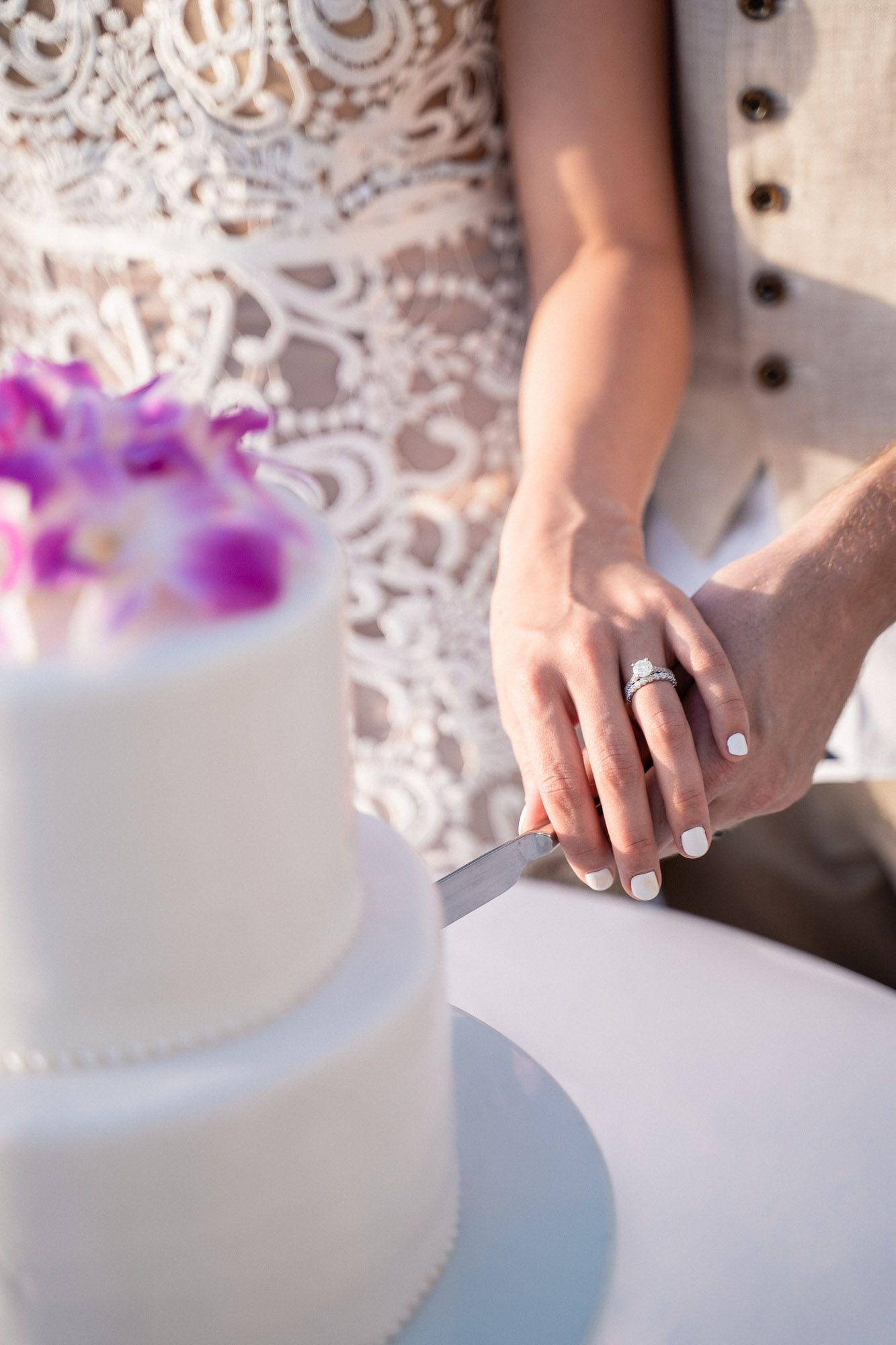 Close-up of wedding rings and hands beside a white wedding cake decorated with purple flowers.