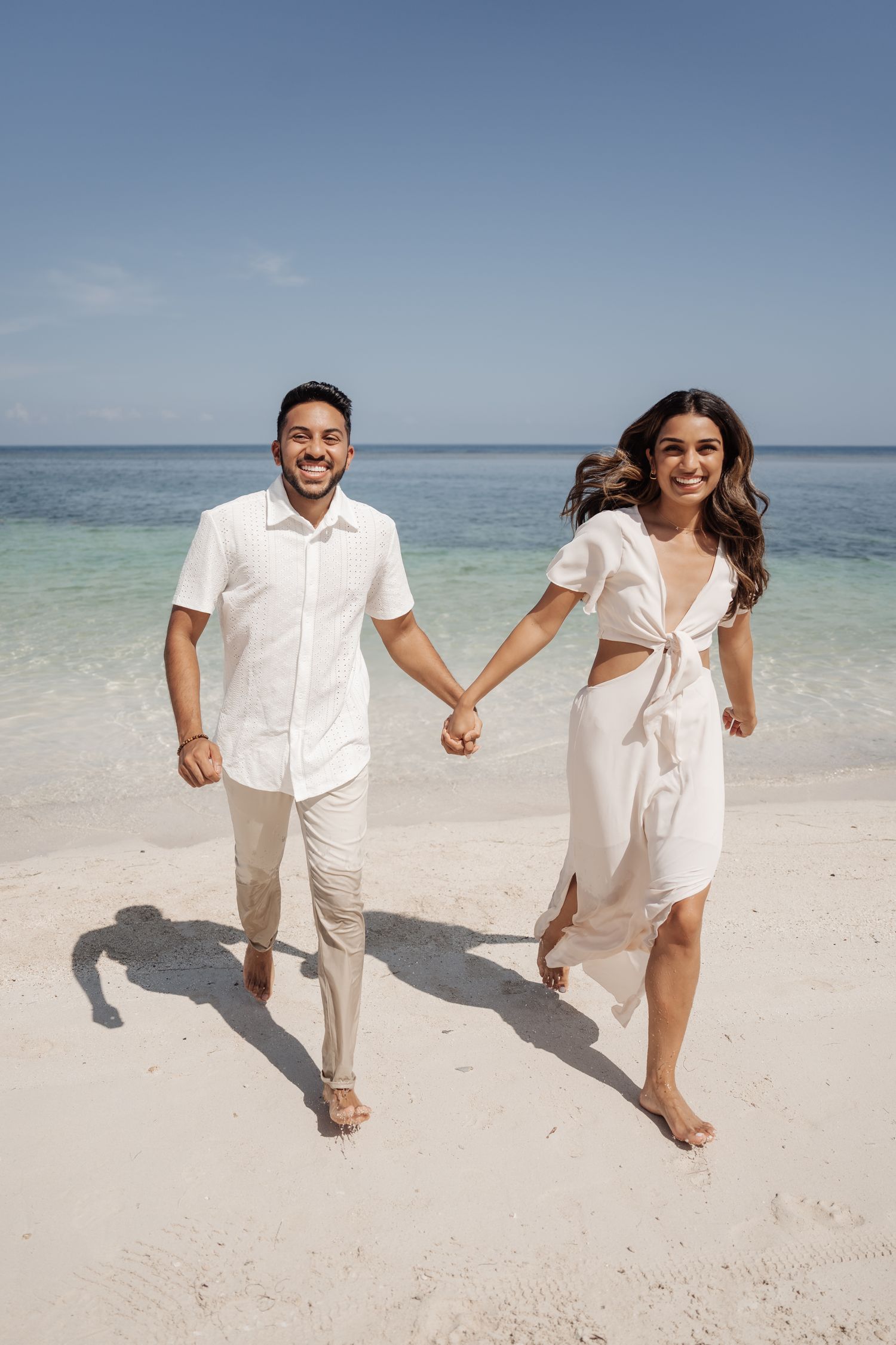 A couple runs joyfully hand in hand along a pristine white sandy beach.