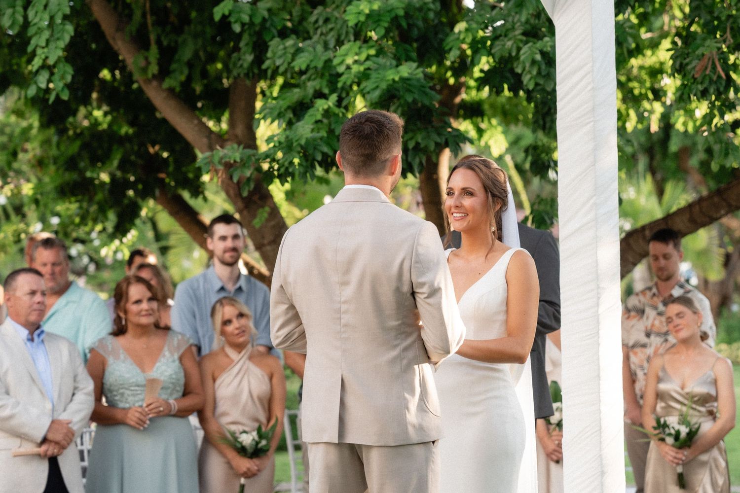 Wedding couple exchanges vows under a lush green canopy while guests look on during an outdoor ceremony.