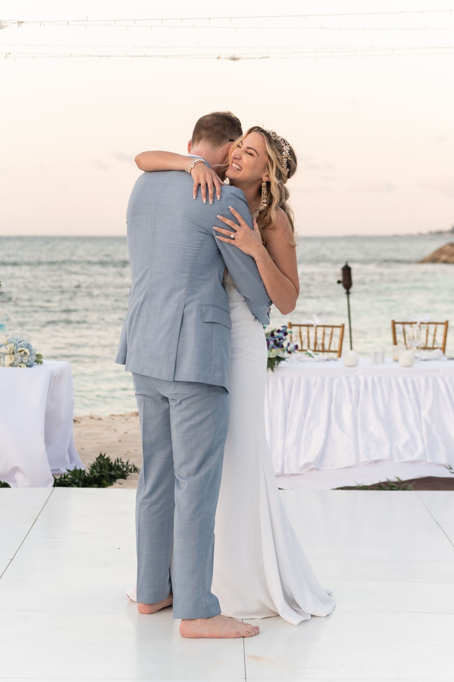 Couple embracing on beach during sunset with reception tables in background.