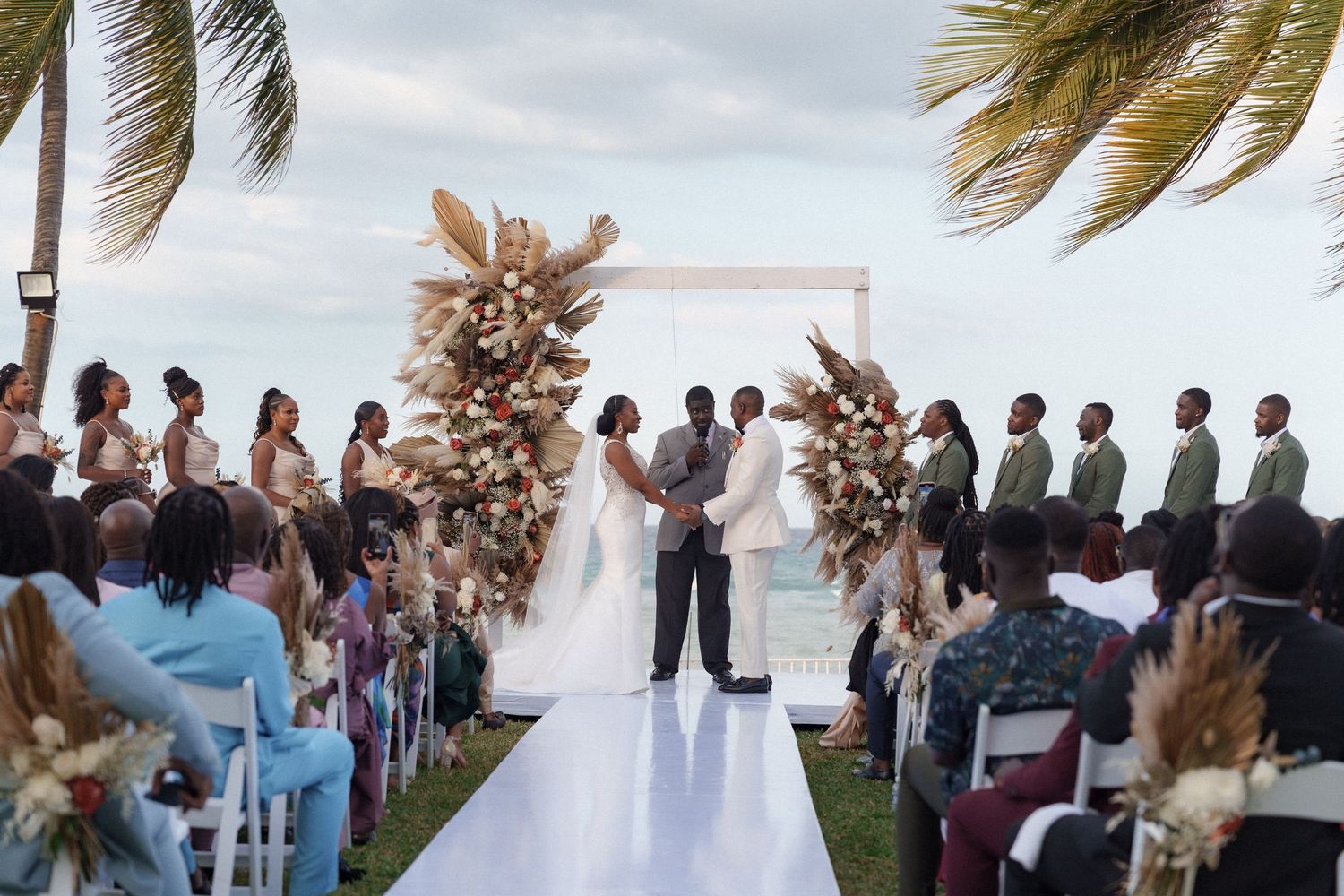 Beachside wedding ceremony with a white aisle runner and floral arch under swaying palm trees.
