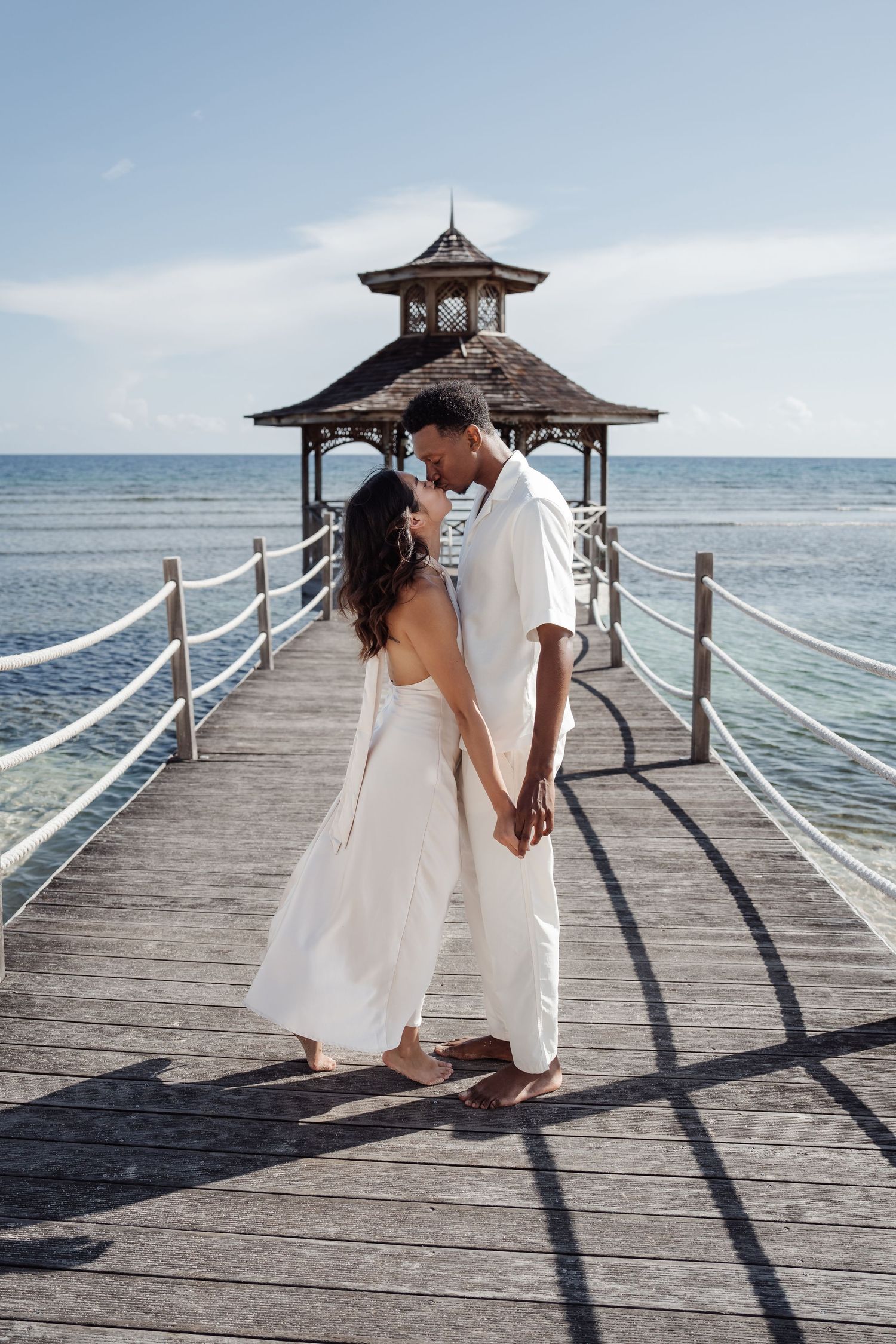A couple shares a romantic moment on a wooden pier with a gazebo in the background.