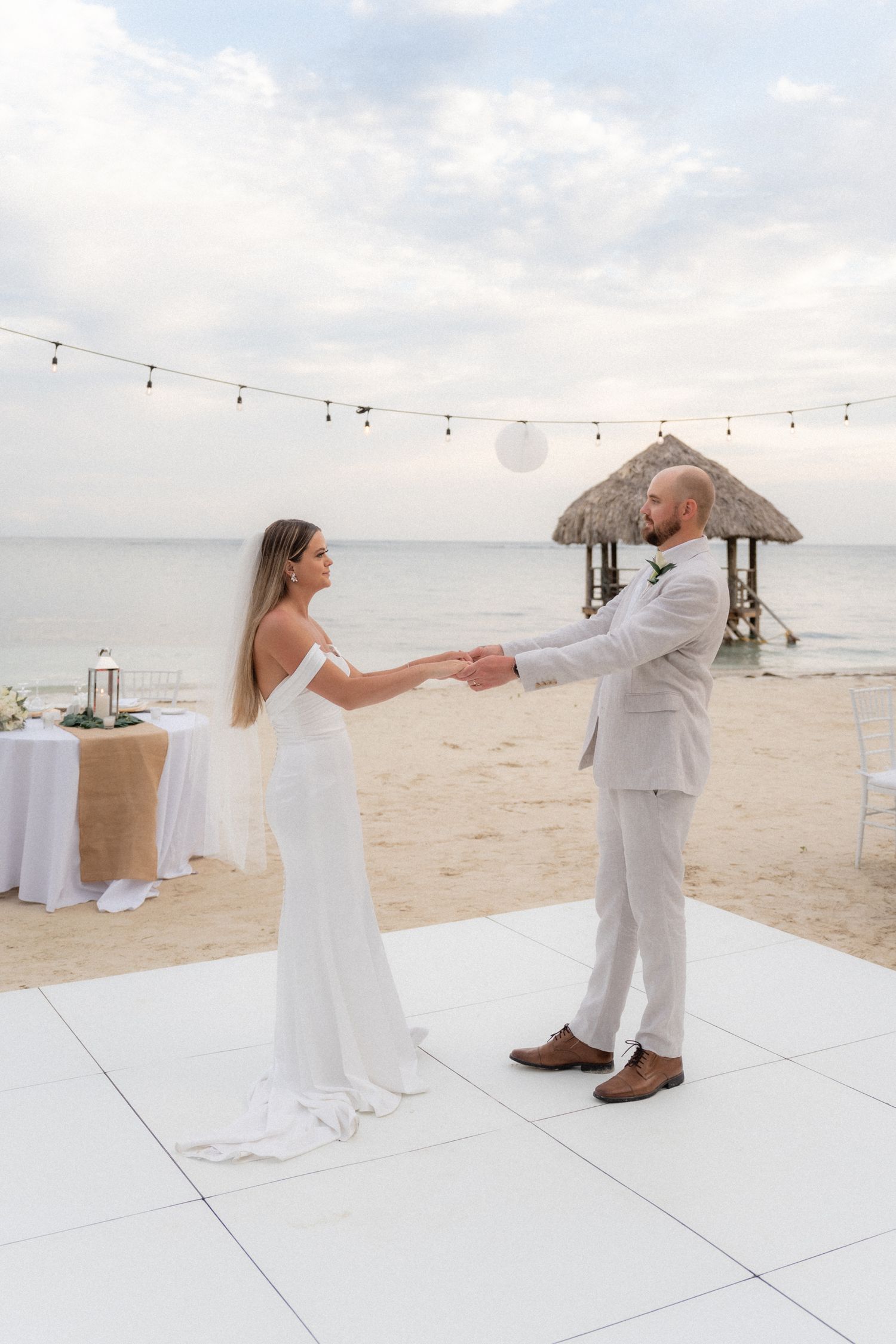 Couple dancing on white platform at beach wedding with string lights and thatched roof palapa in background.